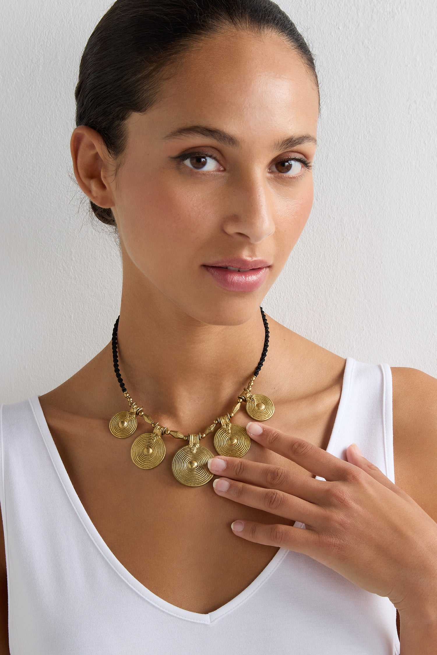 A woman in a white tank top touches the Elissos Engraved Necklace, featuring gold circular pendants, against a plain white background.