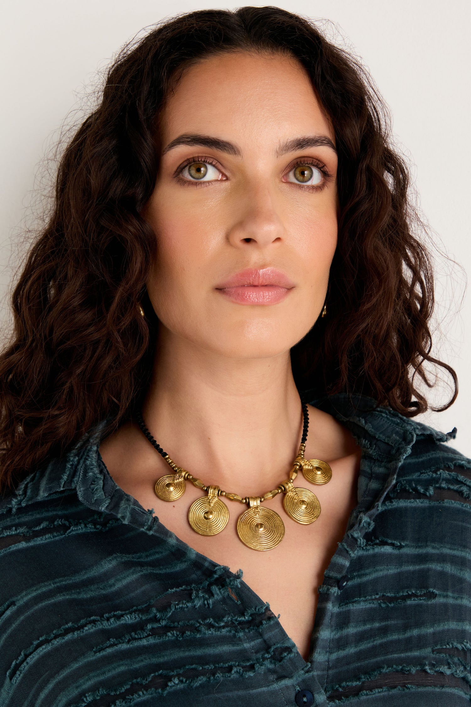 Woman with long curly hair wears a blue textured shirt and the Elissos Engraved Necklace, featuring a bold brass pendant, as she looks slightly upward against a plain background.