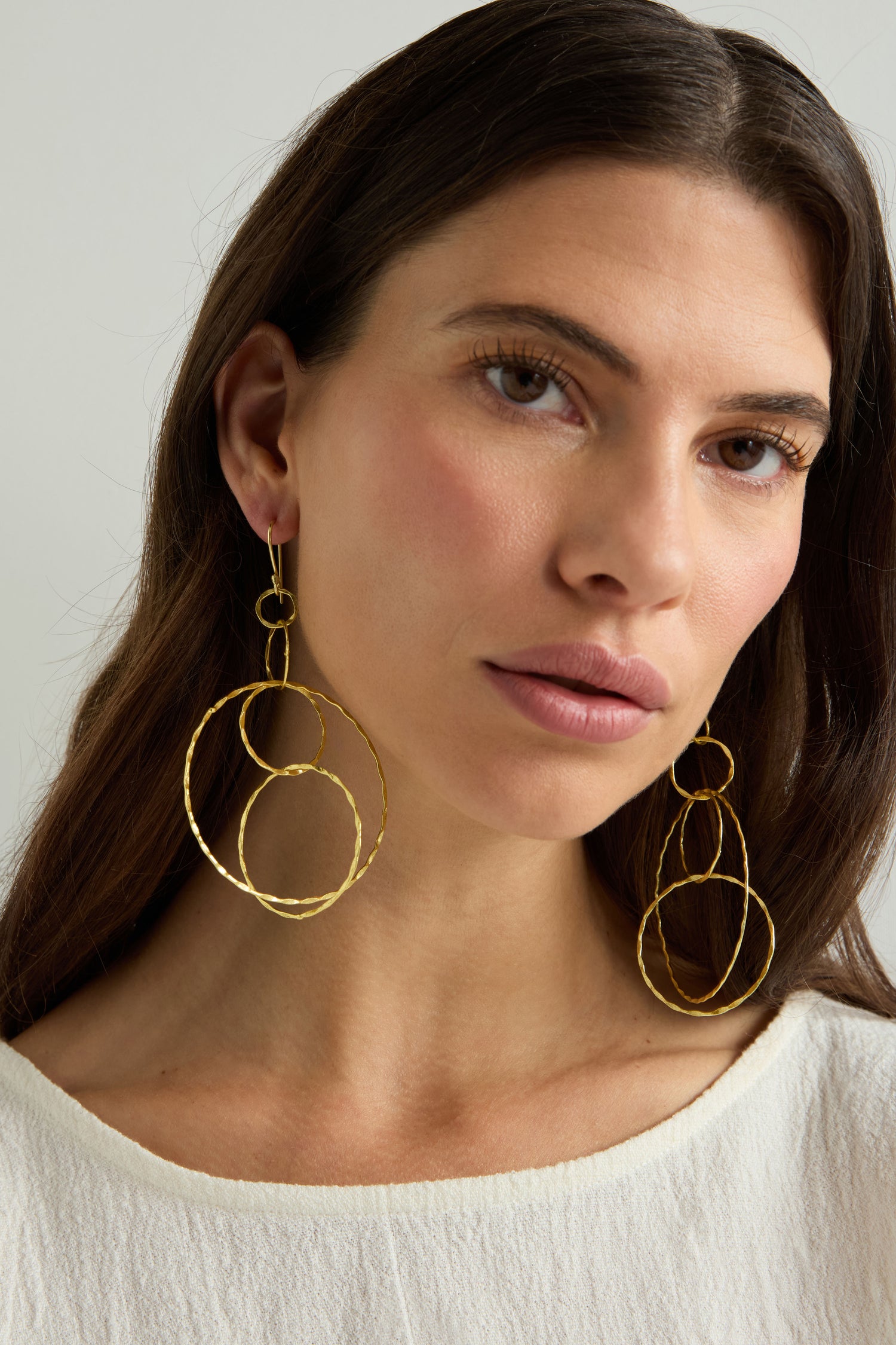 A woman with long brown hair wears Dharma Earrings—stylish gold-finished hoops from the Nataraj Collection—and a white top, gazing slightly to the side against a neutral background.