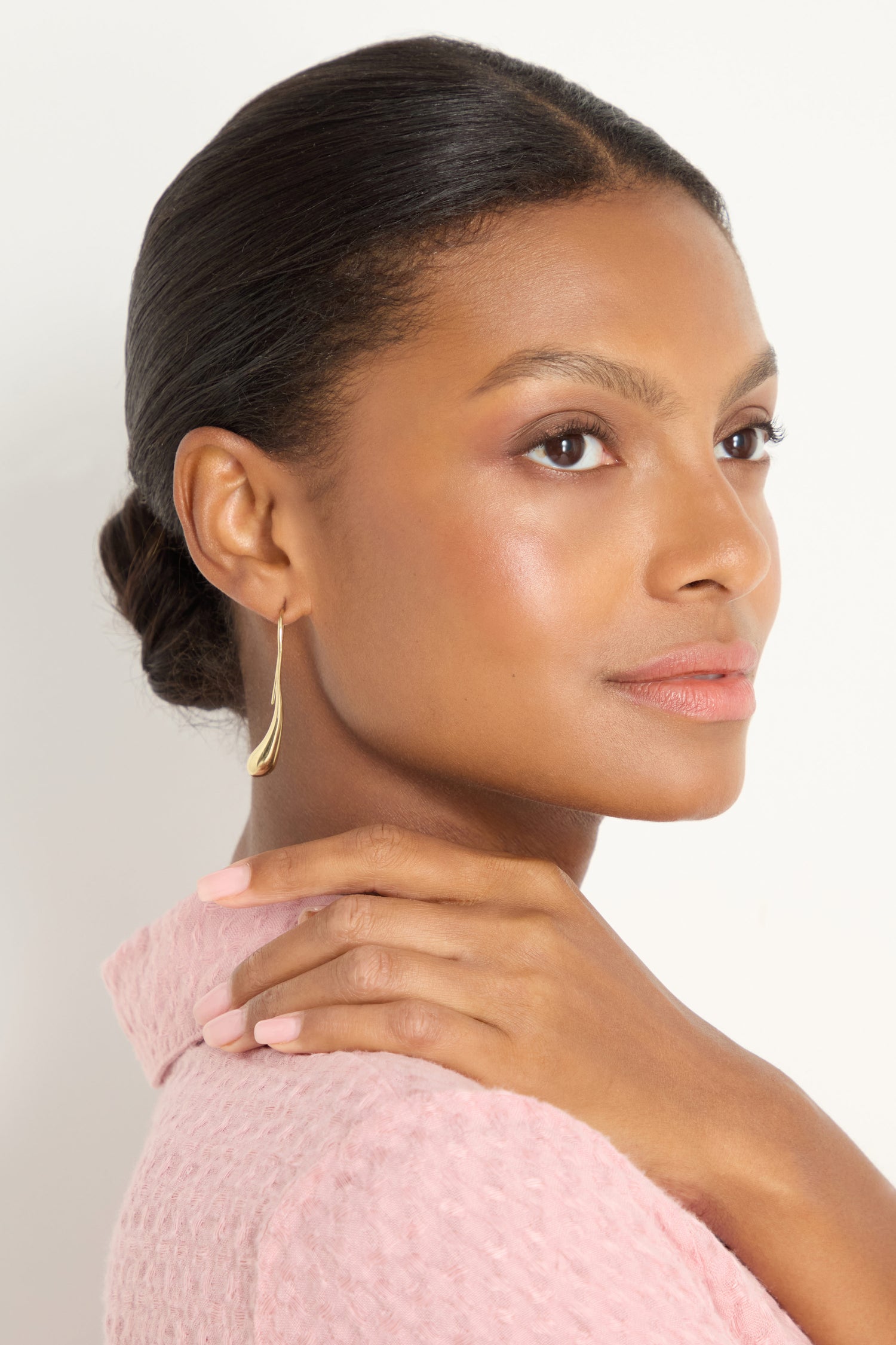 A woman with smooth skin and natural makeup wears Gilded Drop Earrings and a light pink textured top, posing with one hand resting on her shoulder against a plain background.