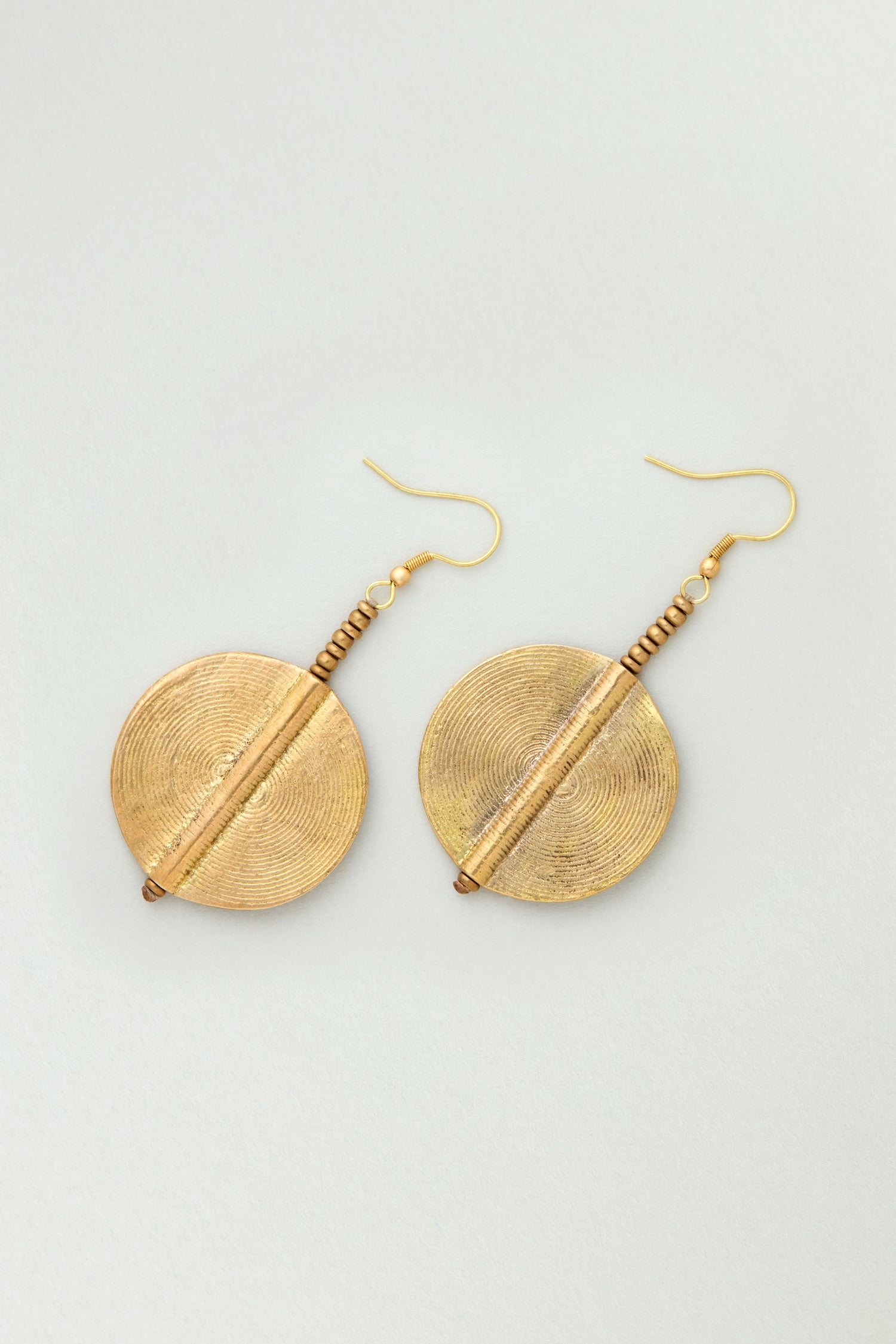 The Spiral Earrings are round, textured brass earrings with hook fastenings, evoking sculptural jewelry style and shown on a light background.