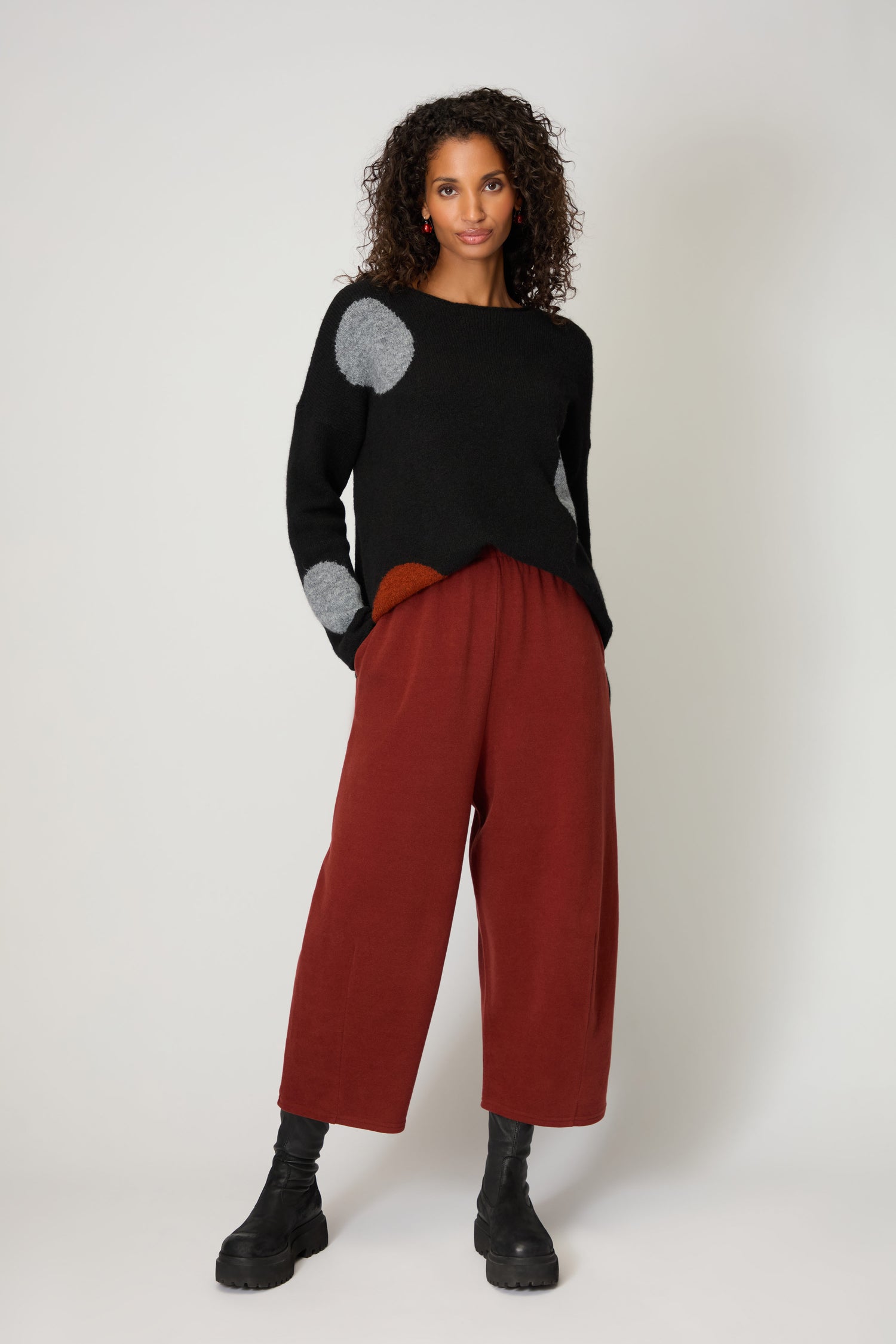 Woman stands against a plain backdrop in Soft Fleece Bubble Trousers (rust), black sweater with circular patches, and black boots—hands in pockets, exuding cosy luxury.