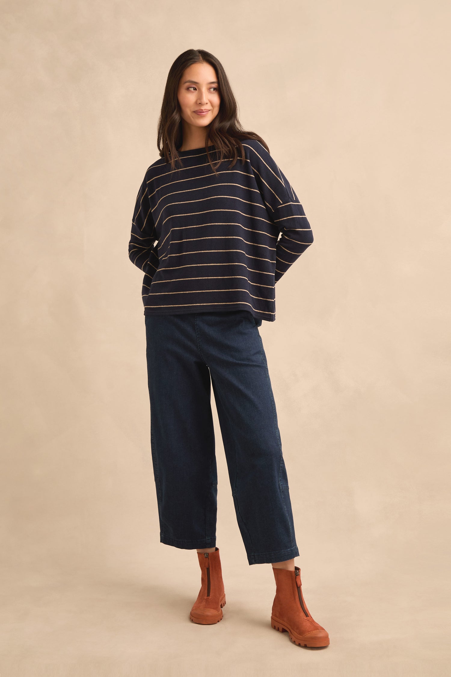 A woman wearing the Narrow Stripe Boxy Top in navy, styled with wide-leg dark blue jeans and brown ankle boots, stands against a beige background.