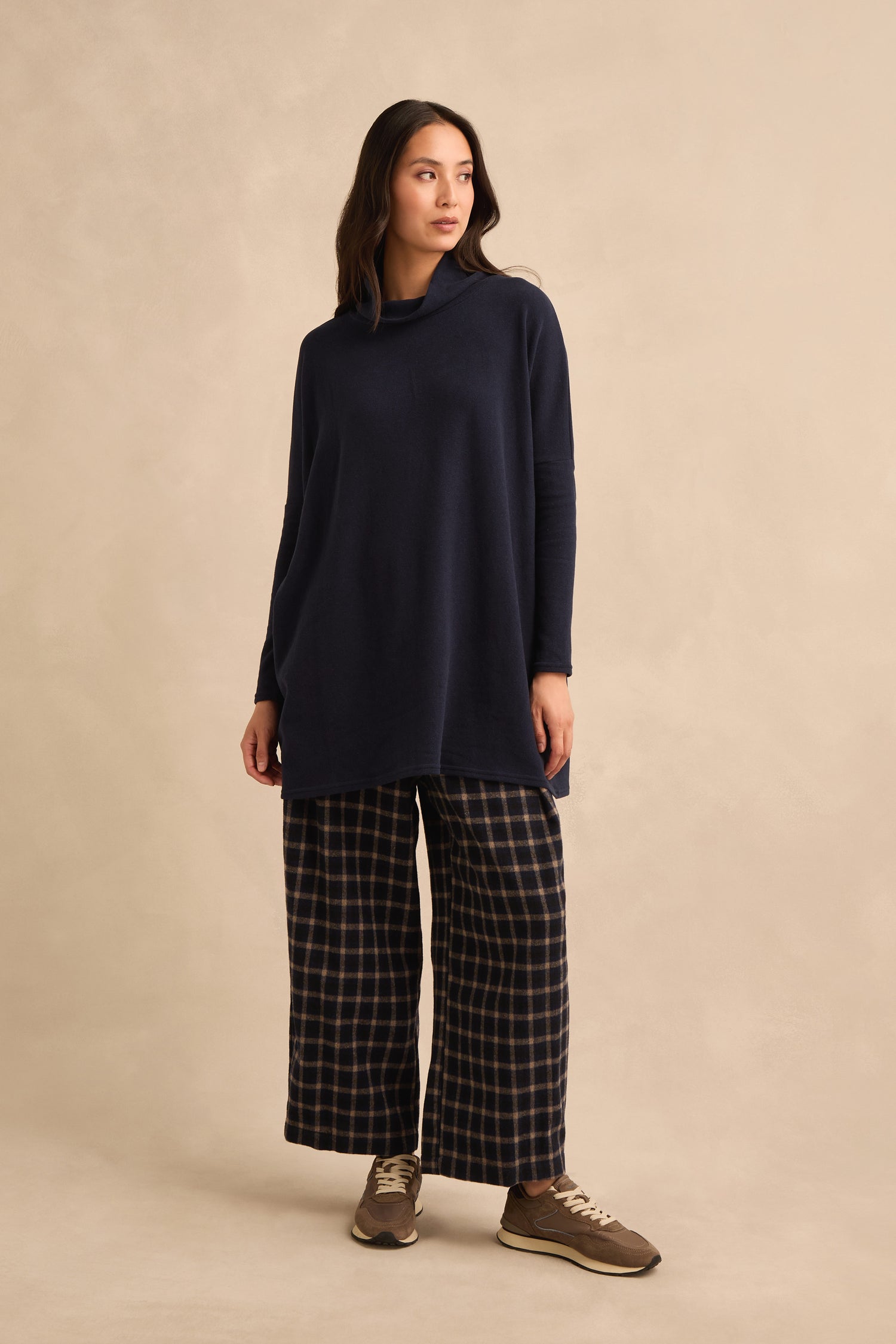 A woman stands against a beige background, wearing a loose Cowl Neck Navy Fleece Tunic, wide-leg plaid pants, and brown sneakers, looking slightly to the side.