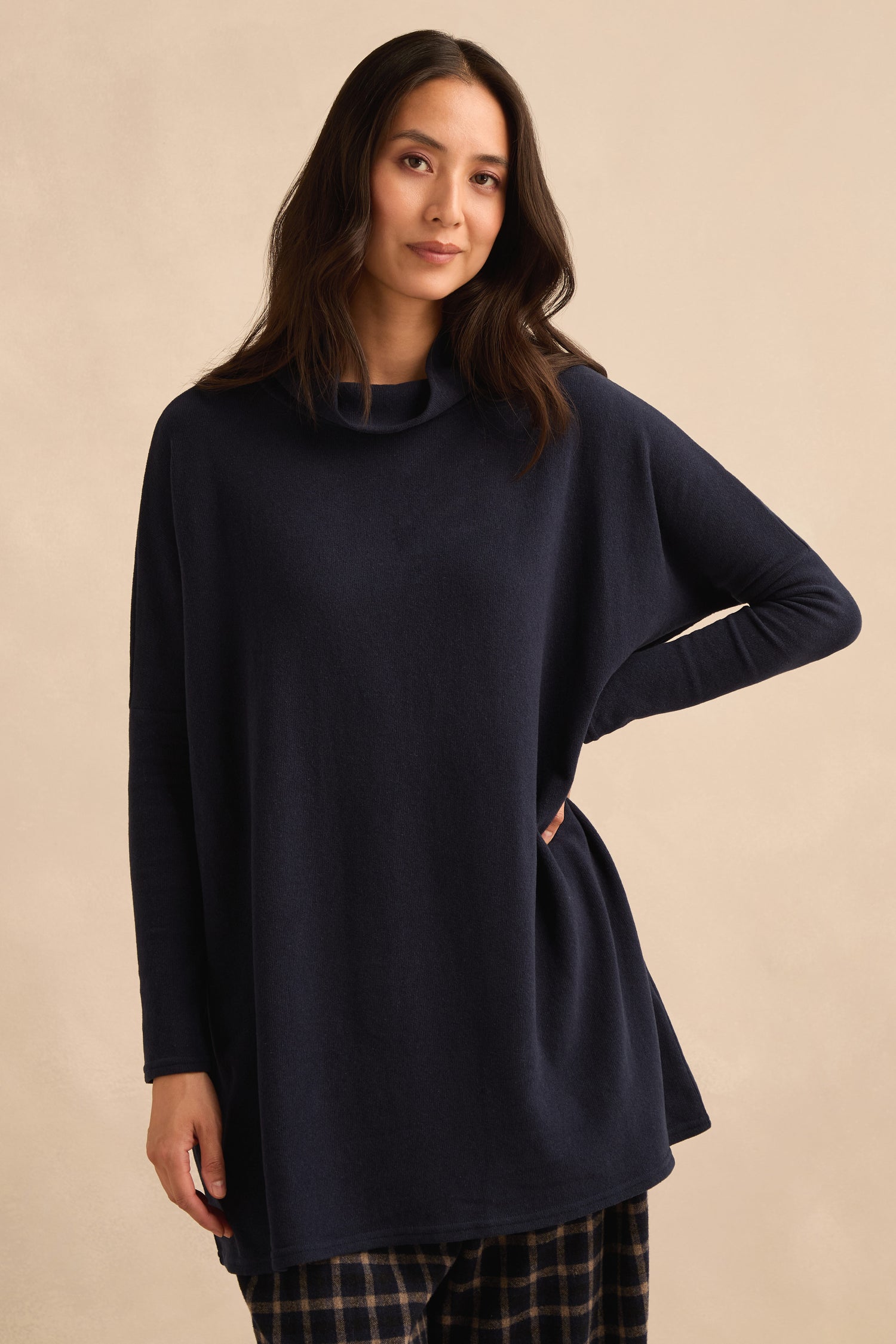 A woman stands against a neutral background with one hand on her hip, wearing the Cowl Neck Navy Fleece Tunic—a loose-fitting, longline sweater made in Italy—paired with plaid pants.