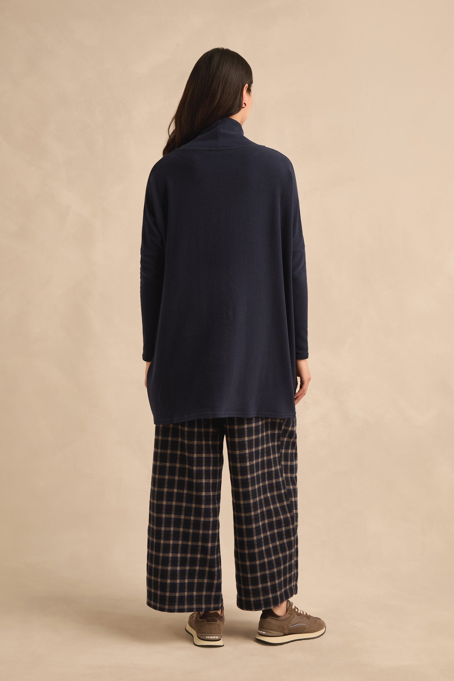 A person stands facing away, wearing the Cowl Neck Navy Fleece Tunic styled as a longline top, paired with wide-legged plaid pants and brown sneakers, against a beige background.