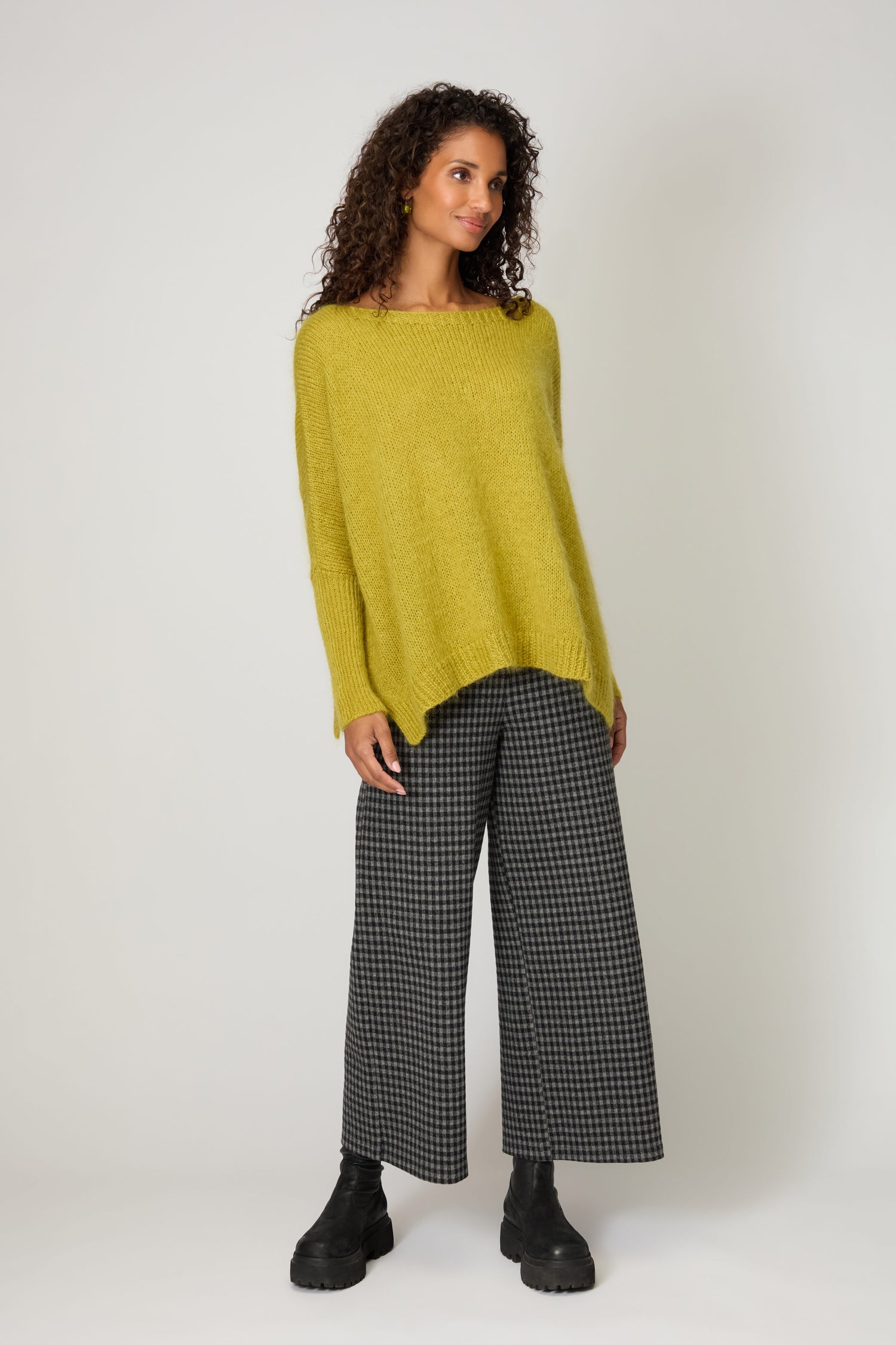 A woman wears the Wide Neck Mohair Knit, an artisan-made, loose yellow sweater from Italy, paired with gray checkered wide-leg pants and black boots, standing against a plain white background.