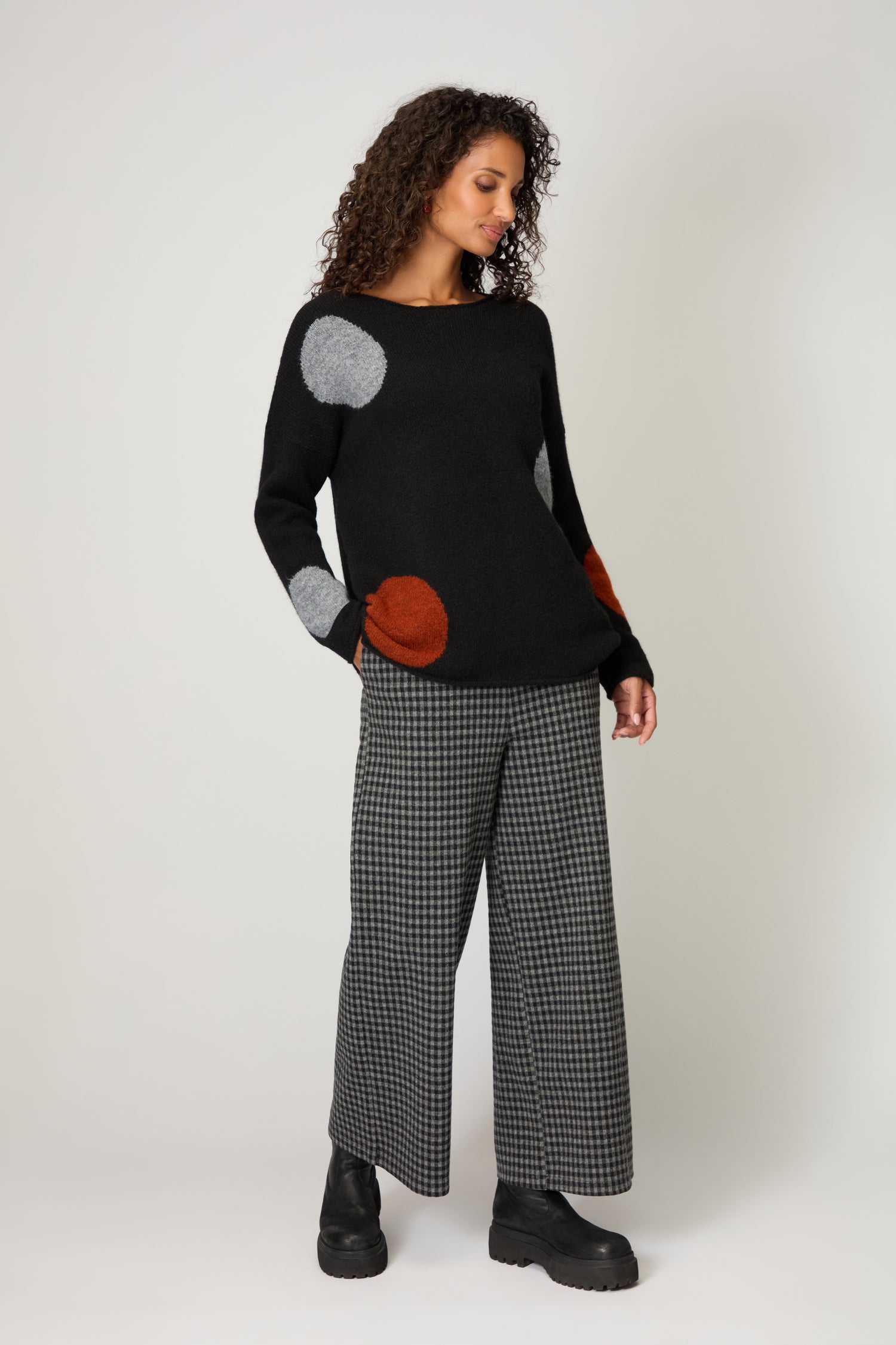 Woman wearing a handmade-in-Italy Multi Spot Knit Top in black with multicolour spots, paired with gray plaid wide-leg pants and black boots, standing against a plain background for a unique and stylish look.