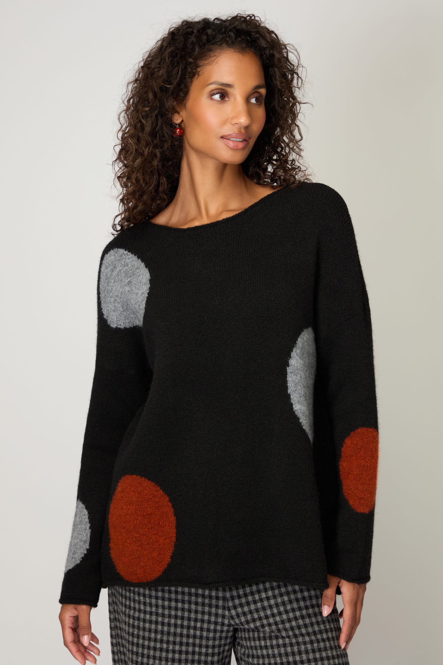 A woman wears the Multi Spot Knit Top—handmade in Italy with large multicolour spots—paired with black and white checkered pants, standing against a plain background.