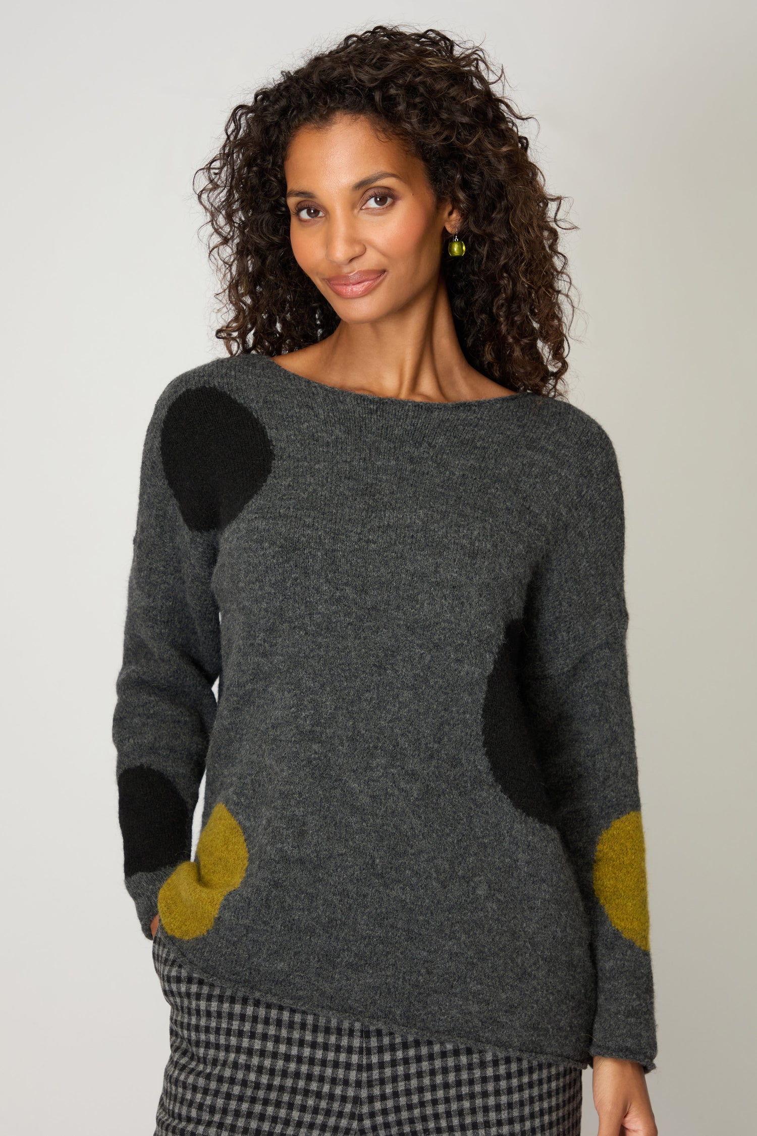 A woman with curly hair wears the Multi Spot Knit Top by Neirami, handmade in Italy, with large black and yellow spots, paired with checkered pants against a plain background.