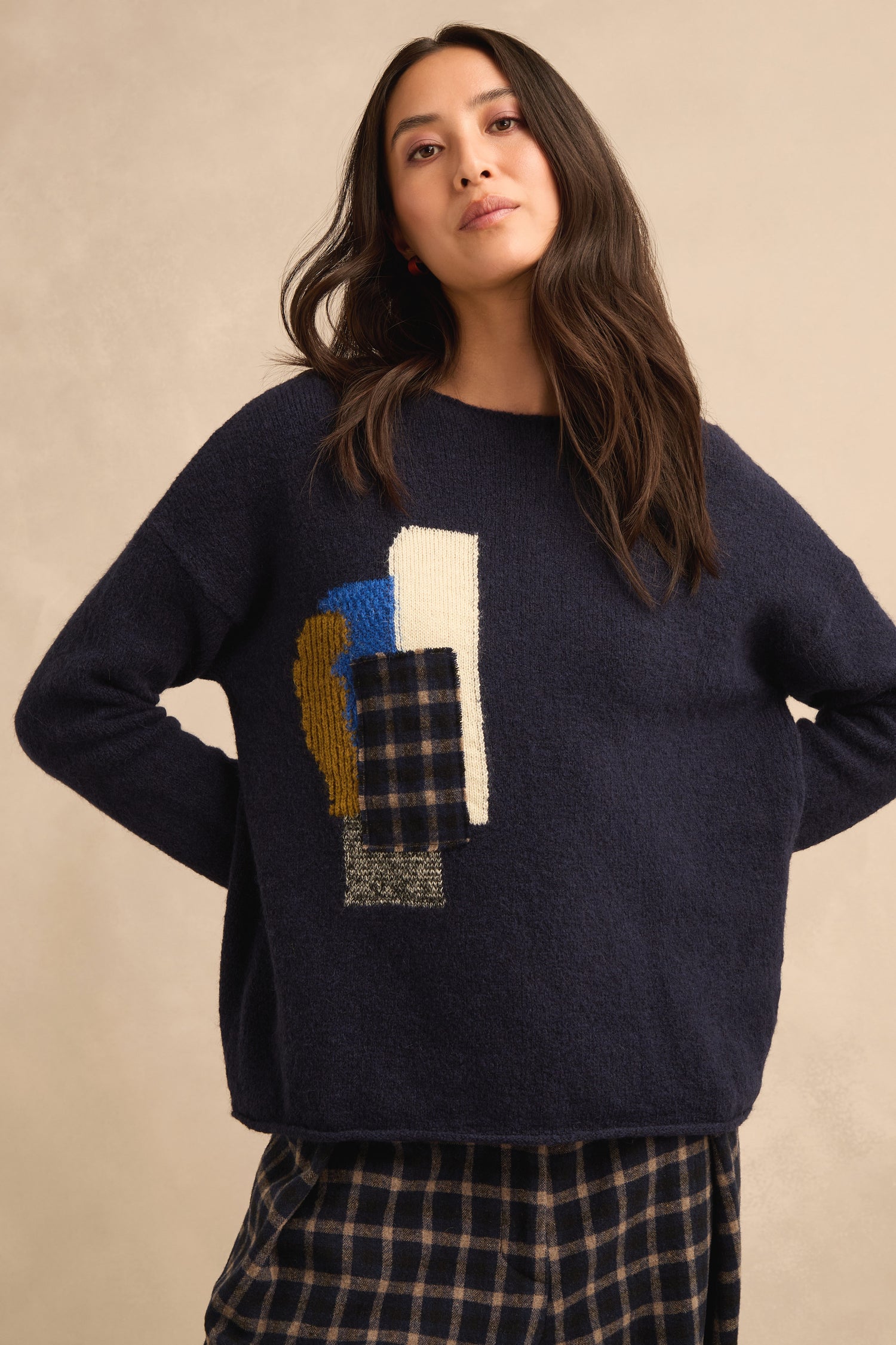 A woman wearing the Patch Detail Jumper—a relaxed fit navy knit with abstract detailing—paired with a navy plaid skirt, stands against a beige background.