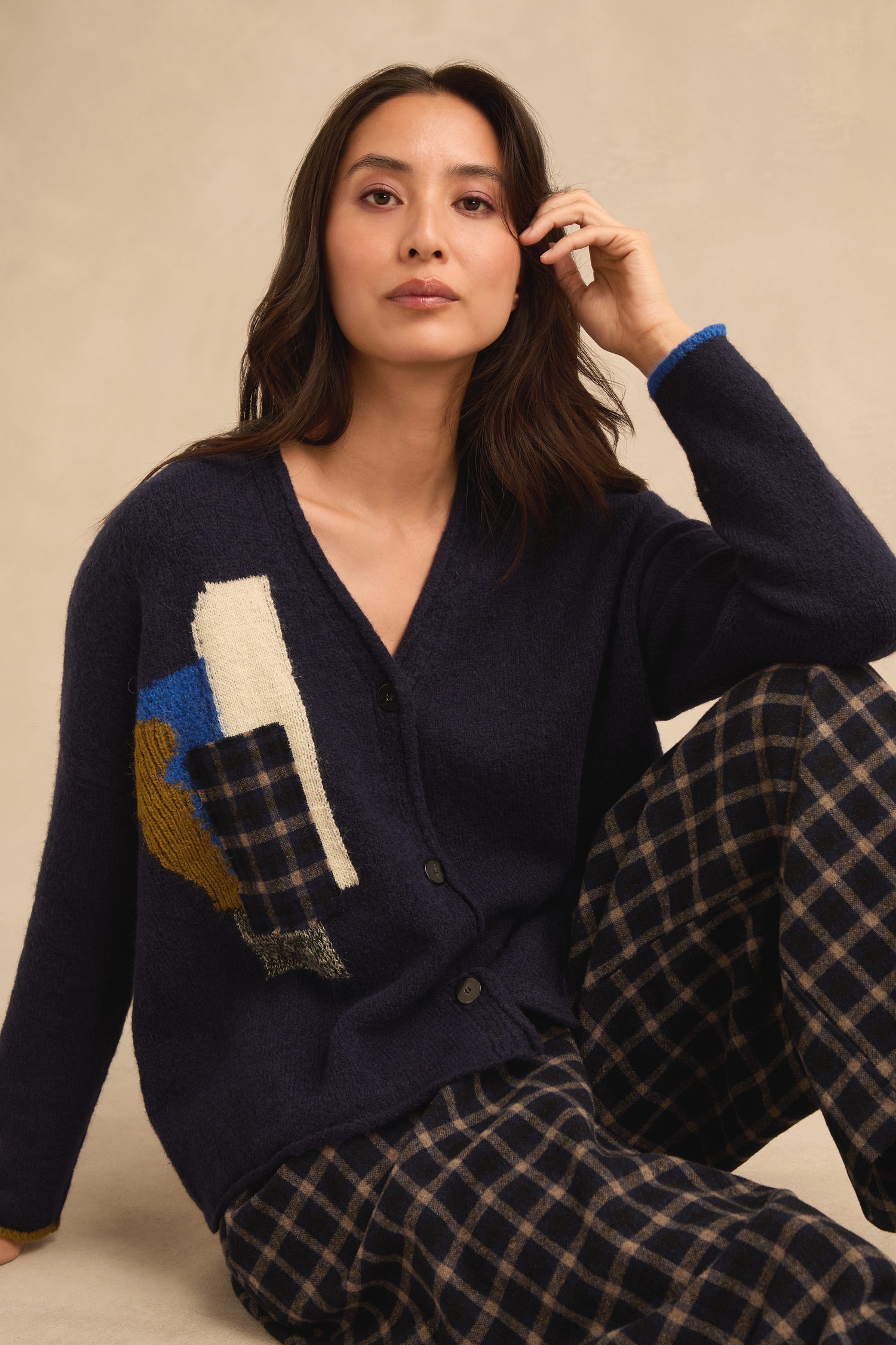 A woman is seated against a neutral background, wearing the Patch Detail Cardigan in navy with abstract patterns and plaid pants. Made in Italy, this stylish cardigan features patch details that showcase its sophisticated design and quality craftsmanship.