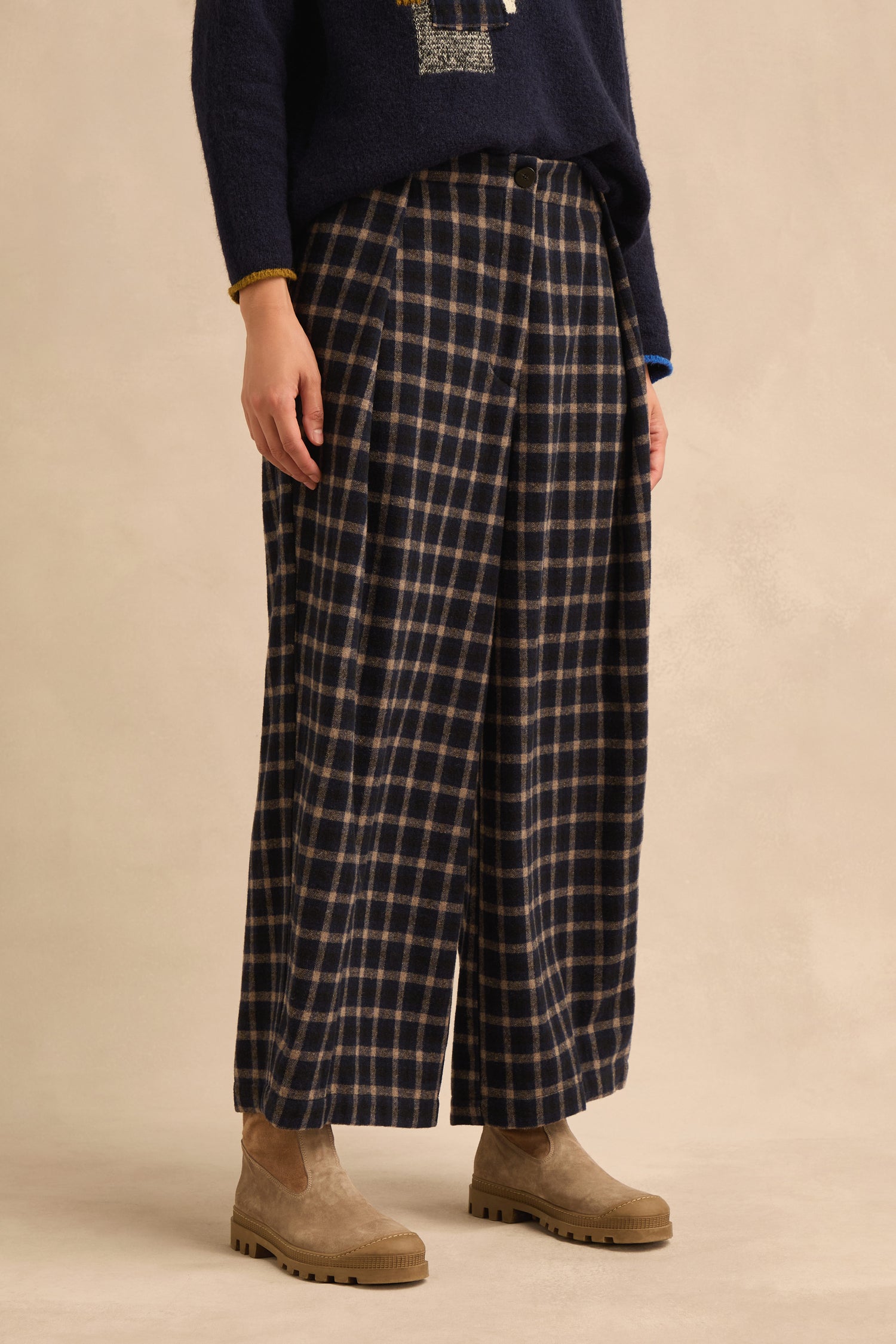 Wearing the Tartan Pleated Trousers with a dark sweater and tan suede boots, a person stands against a plain beige background.