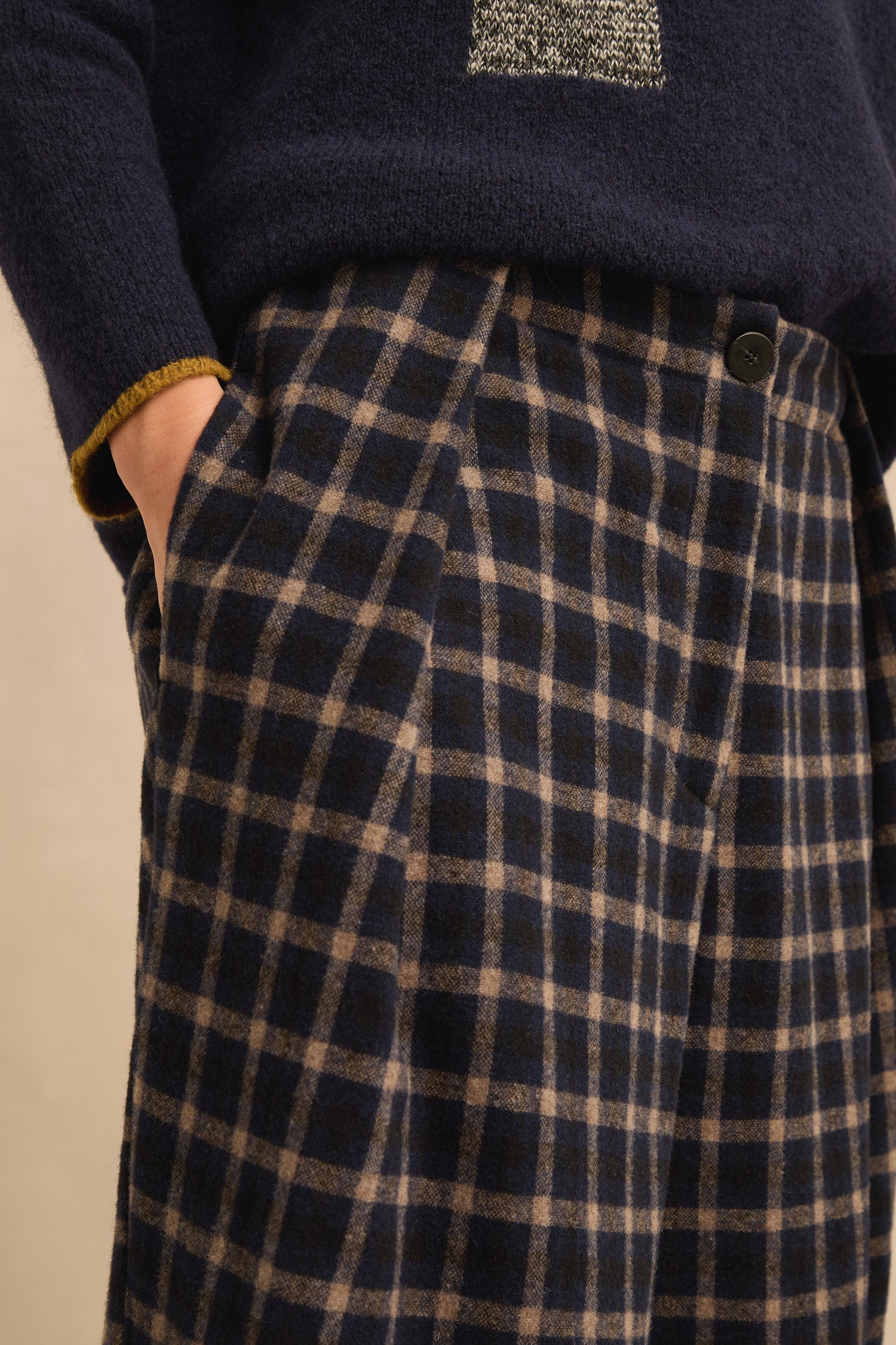 Model is shown from chest to knee, wearing Tartan Pleated Trousers with hands in pockets and a navy blue sweater.