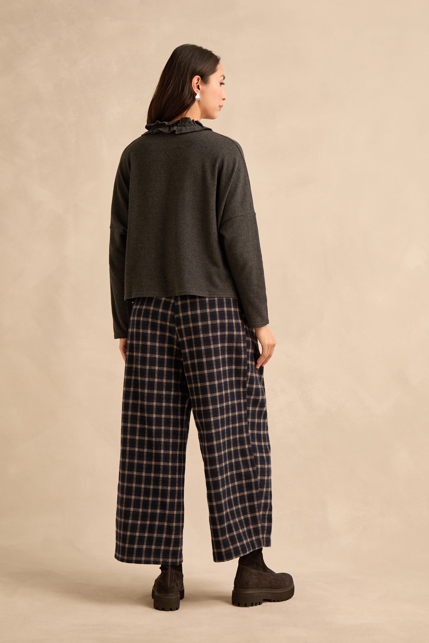 A person stands facing away, wearing a Frill Neck Dark Grey Fleece Cardigan, plaid wide-leg pants, and black boots against a plain beige background.