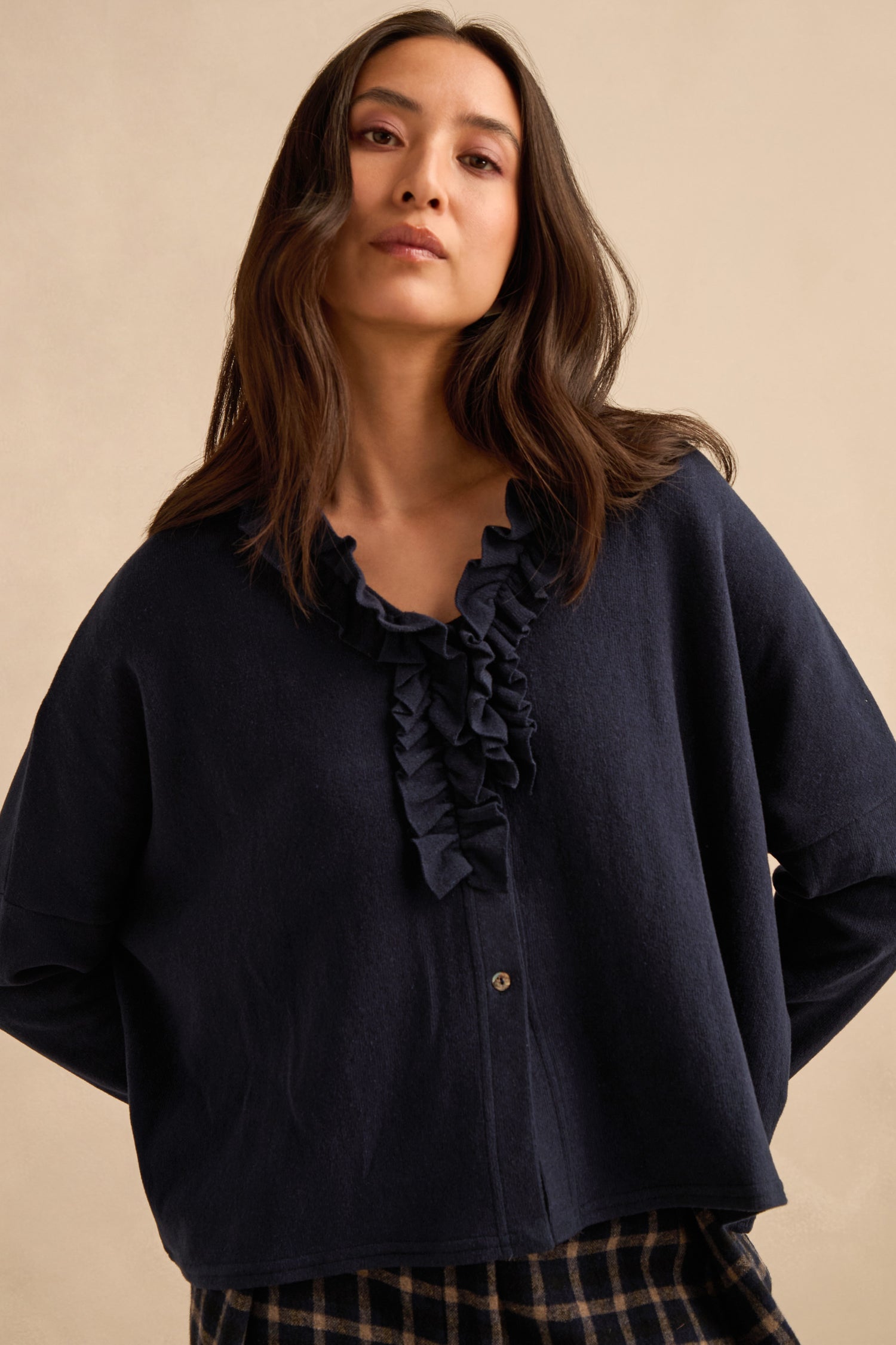 Wearing the Frill Neck Navy Fleece Cardigan layered over a ruffled blouse and paired with plaid pants, the woman stands against a beige background, gazing slightly downward with a relaxed posture.