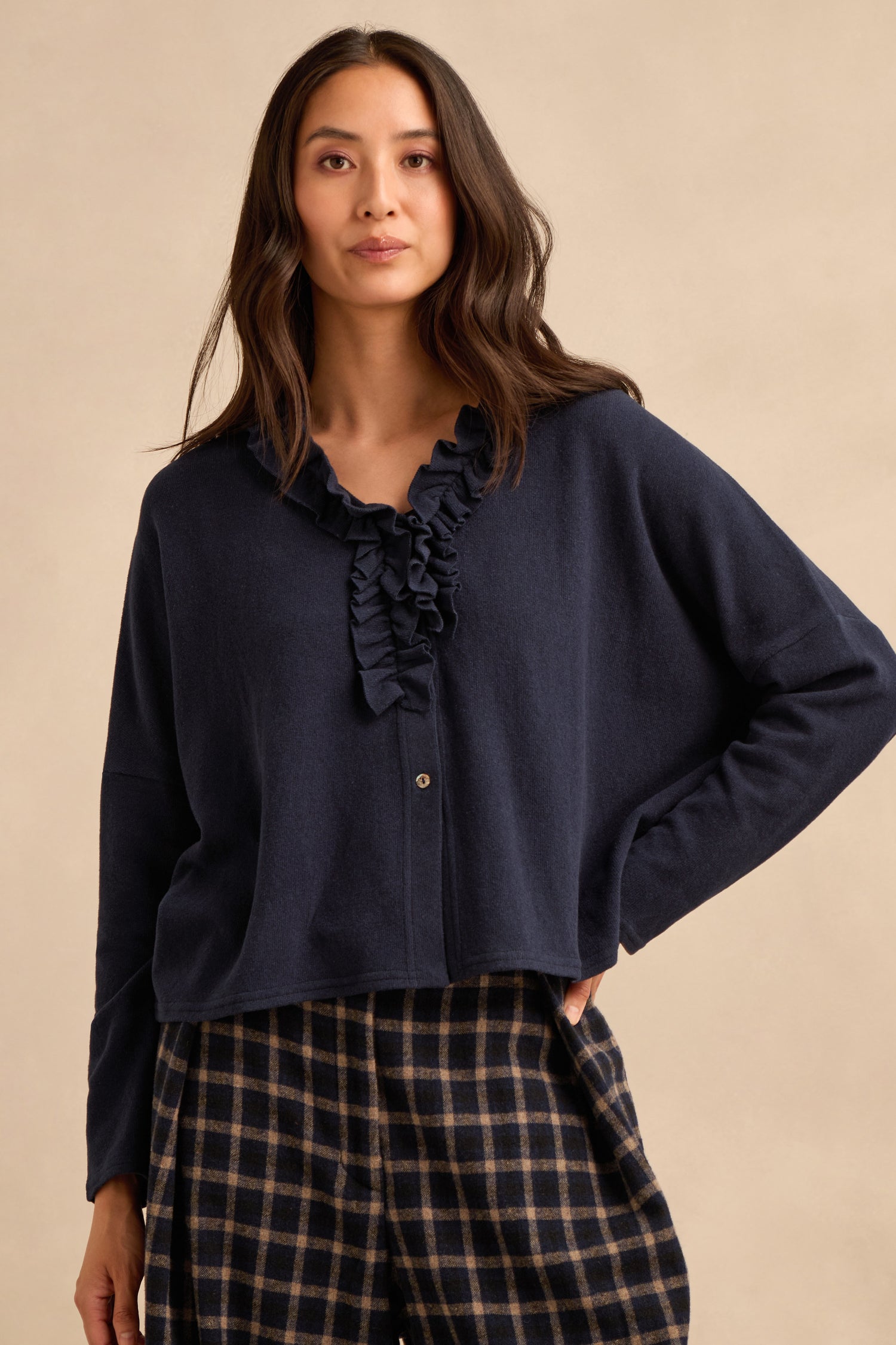 A woman stands in front of a plain beige background, wearing the Frill Neck Navy Fleece Cardigan layered over a navy blouse with a ruffled collar and plaid pants.