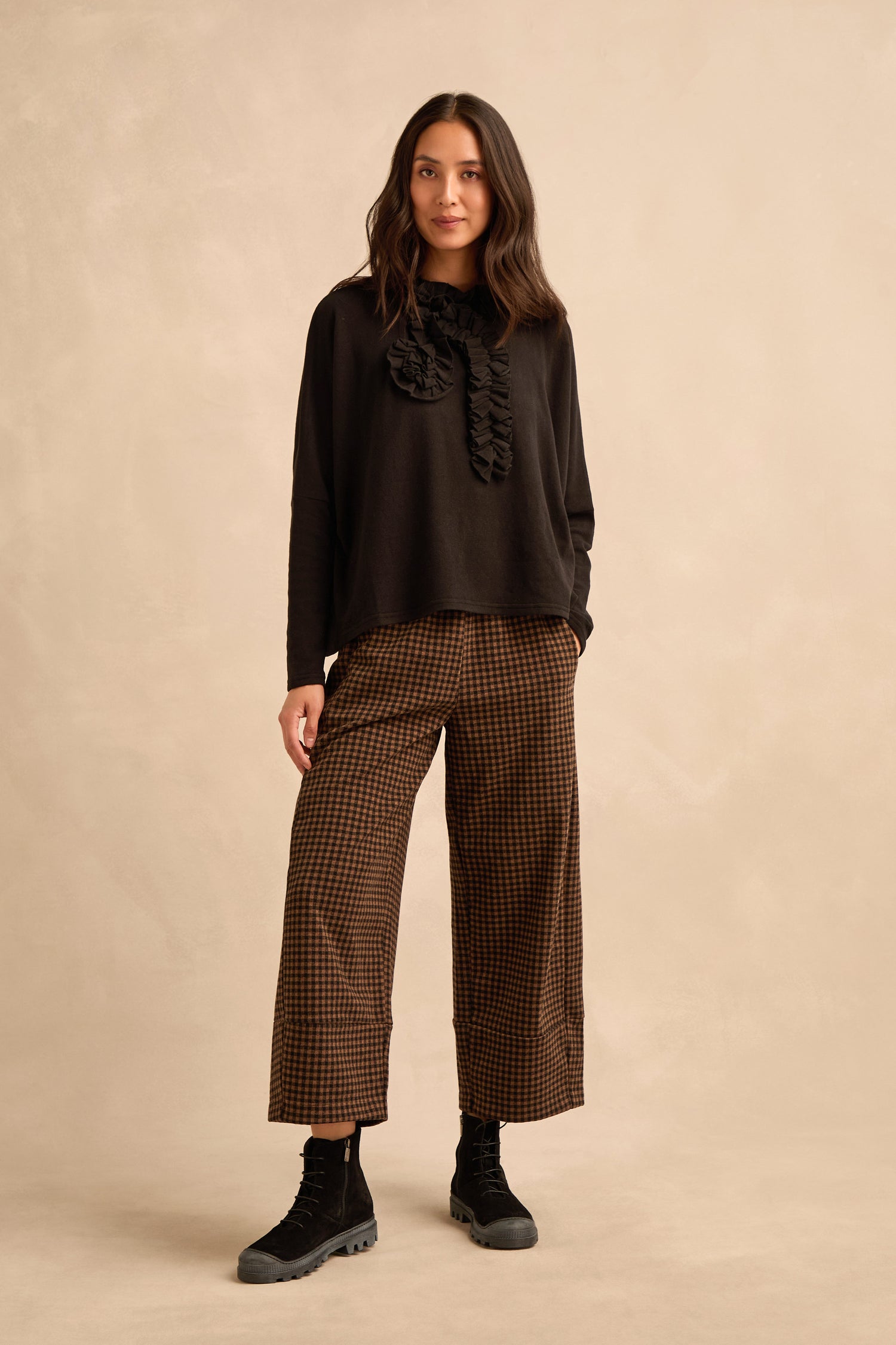 A woman stands against a beige backdrop, wearing the Black Frill Neck Top with ruffle details, brown plaid wide-leg pants, and black lace-up boots.