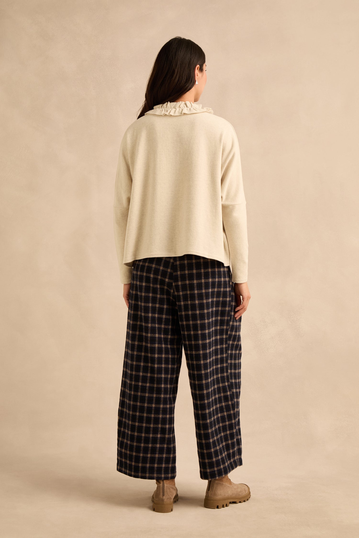 A person stands facing away, wearing the Cream Frill Neck Top with a relaxed boxy fit, paired with wide-leg navy plaid pants and tan boots against a plain beige background.
