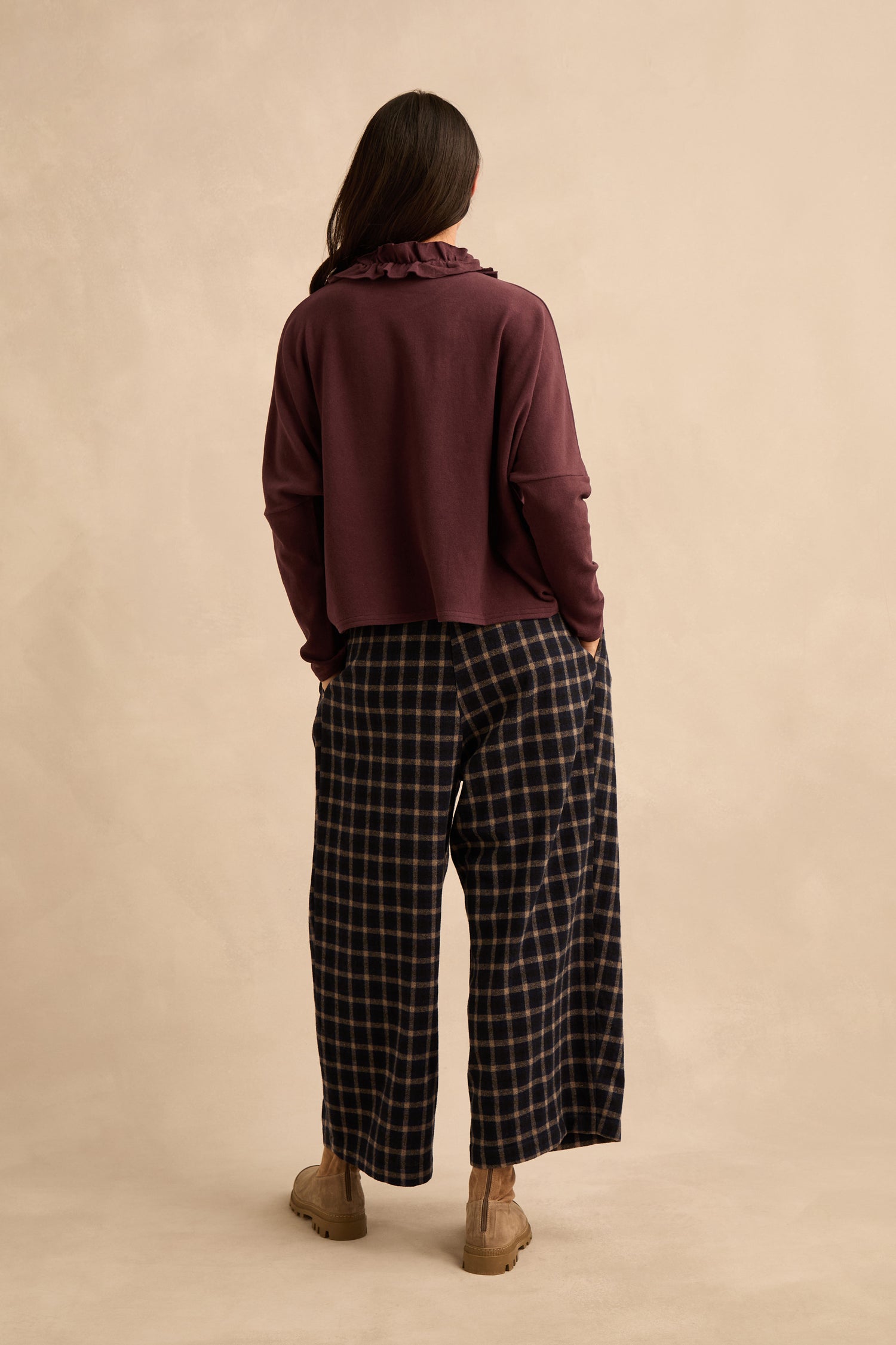 Person standing with back to camera, wearing the Fig Frill Neck Top—a maroon frill neck top with a ruffled collar—paired with wide-leg navy plaid pants and tan shoes, against a plain beige background.