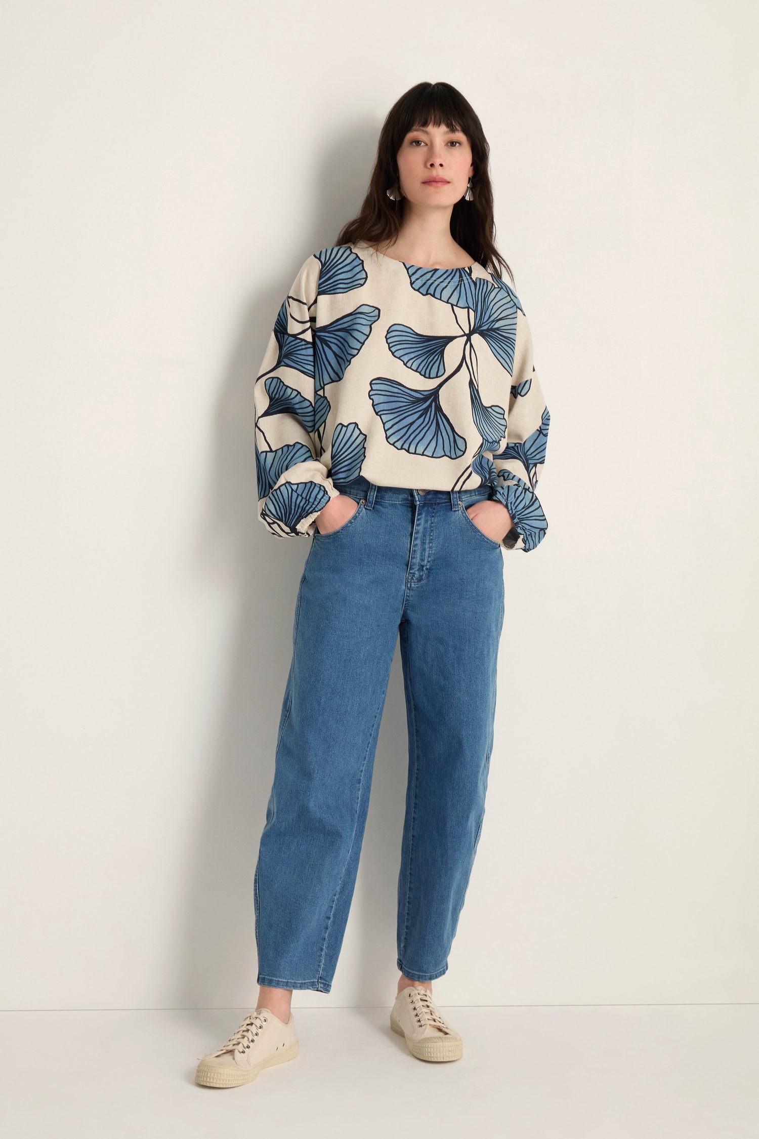 A woman stands against a plain wall, wearing a cream sweater with blue leaf patterns, Stretch Denim Jeans with a five-pocket design that graze the ankles, and beige sneakers.