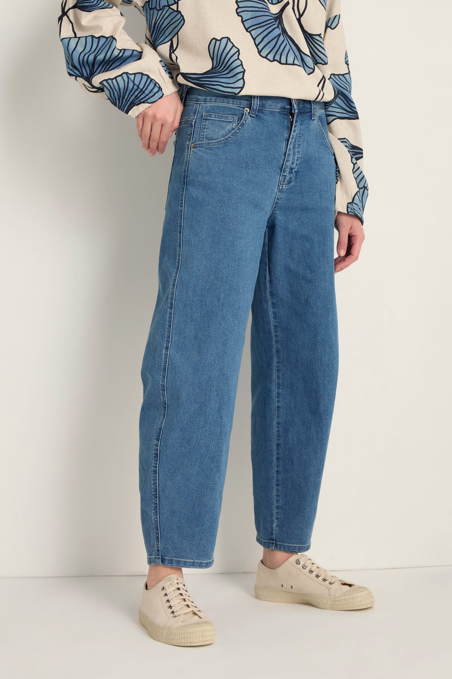 A person wears Stretch Denim Jeans in blue, featuring a five-pocket design and ankle-grazing length, paired with a beige sweater with blue leaf patterns and beige sneakers, standing against a light background.