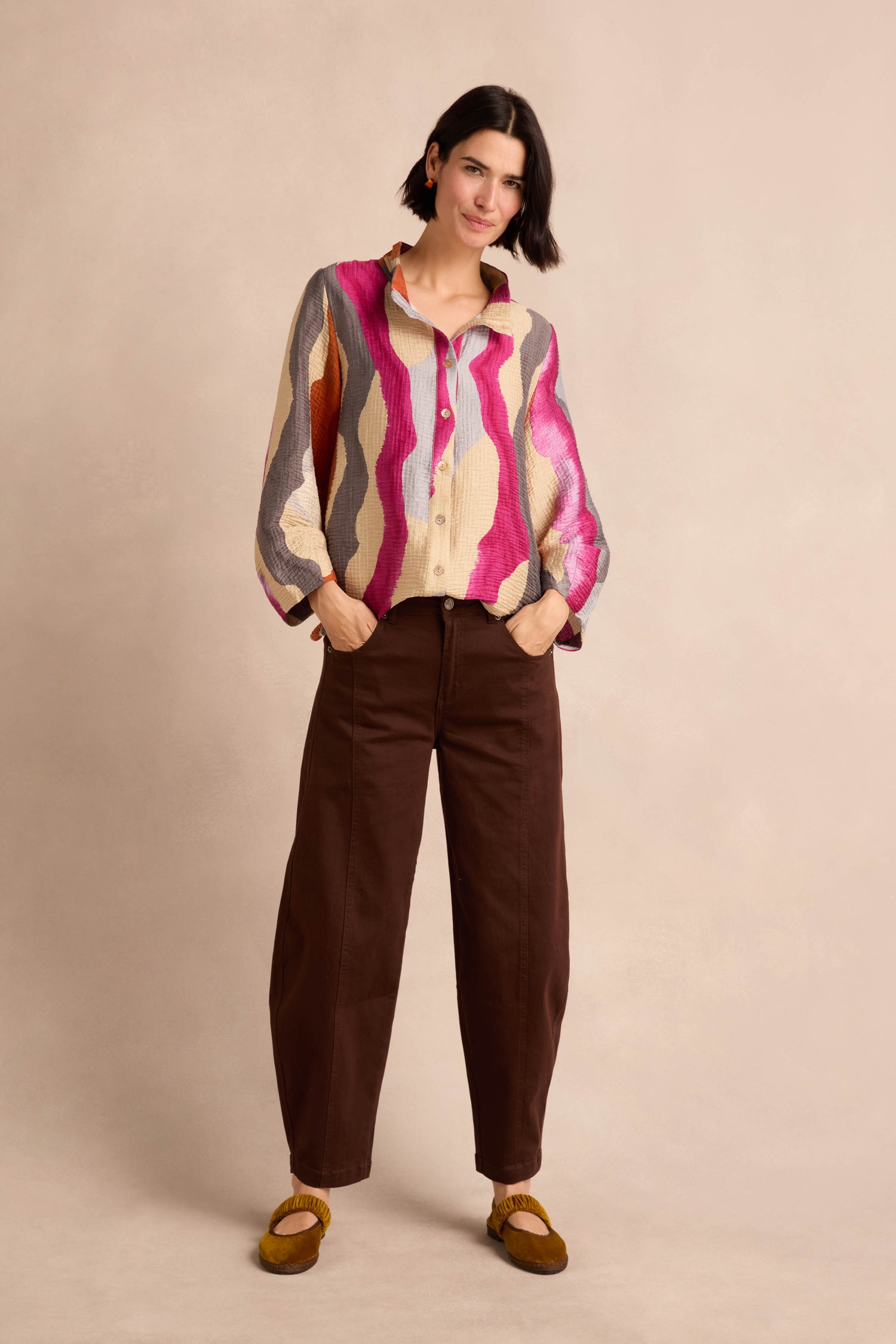 A person stands with their hands in their pockets, wearing a colorful patterned shirt, Stretch Cotton Trousers in a relaxed brown fit, and mustard yellow shoes against a plain beige background.