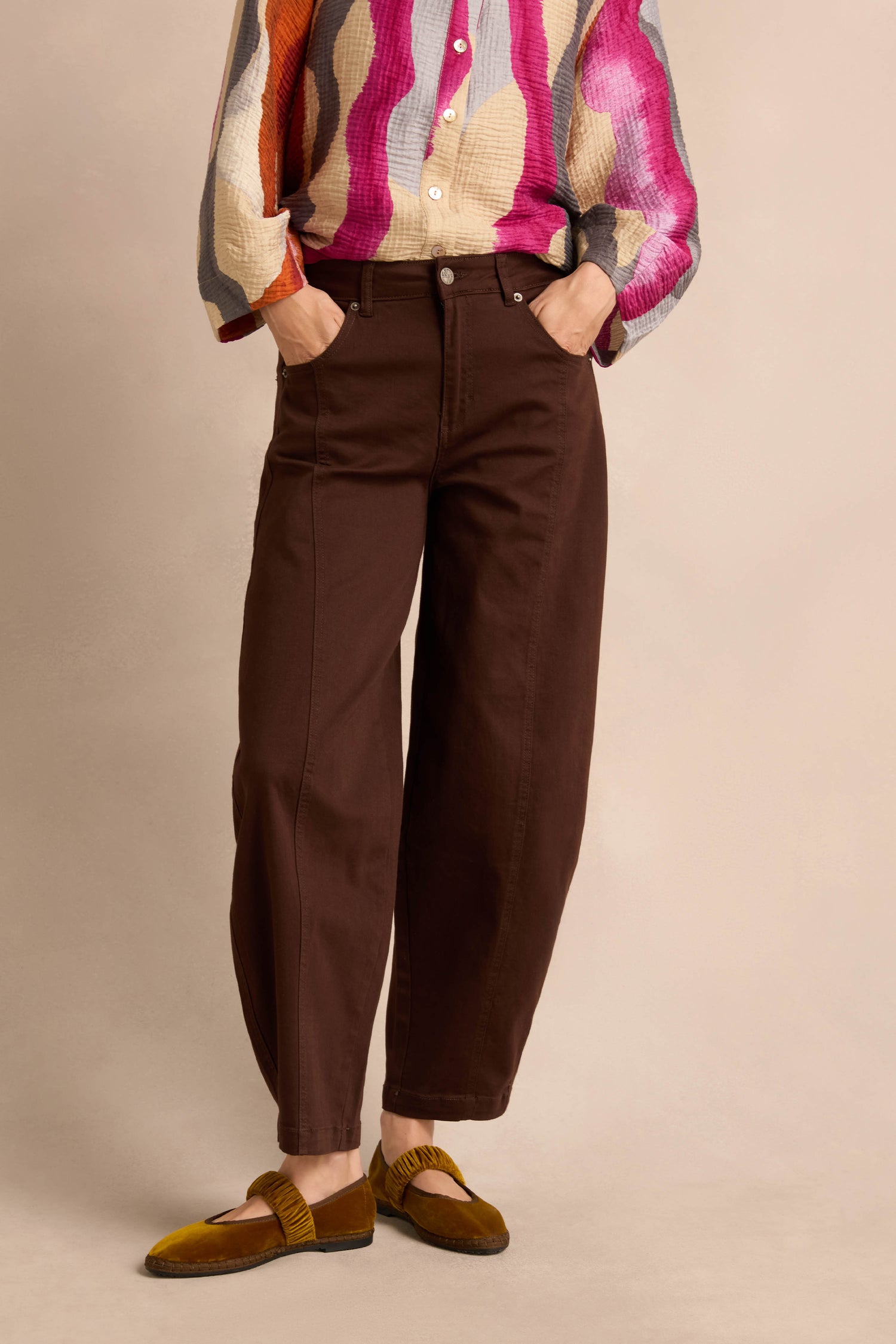 A person wearing relaxed fit Stretch Cotton Trousers in brown, paired with a colorful long-sleeve shirt and brown loafers, stands with hands in pockets against a neutral background.