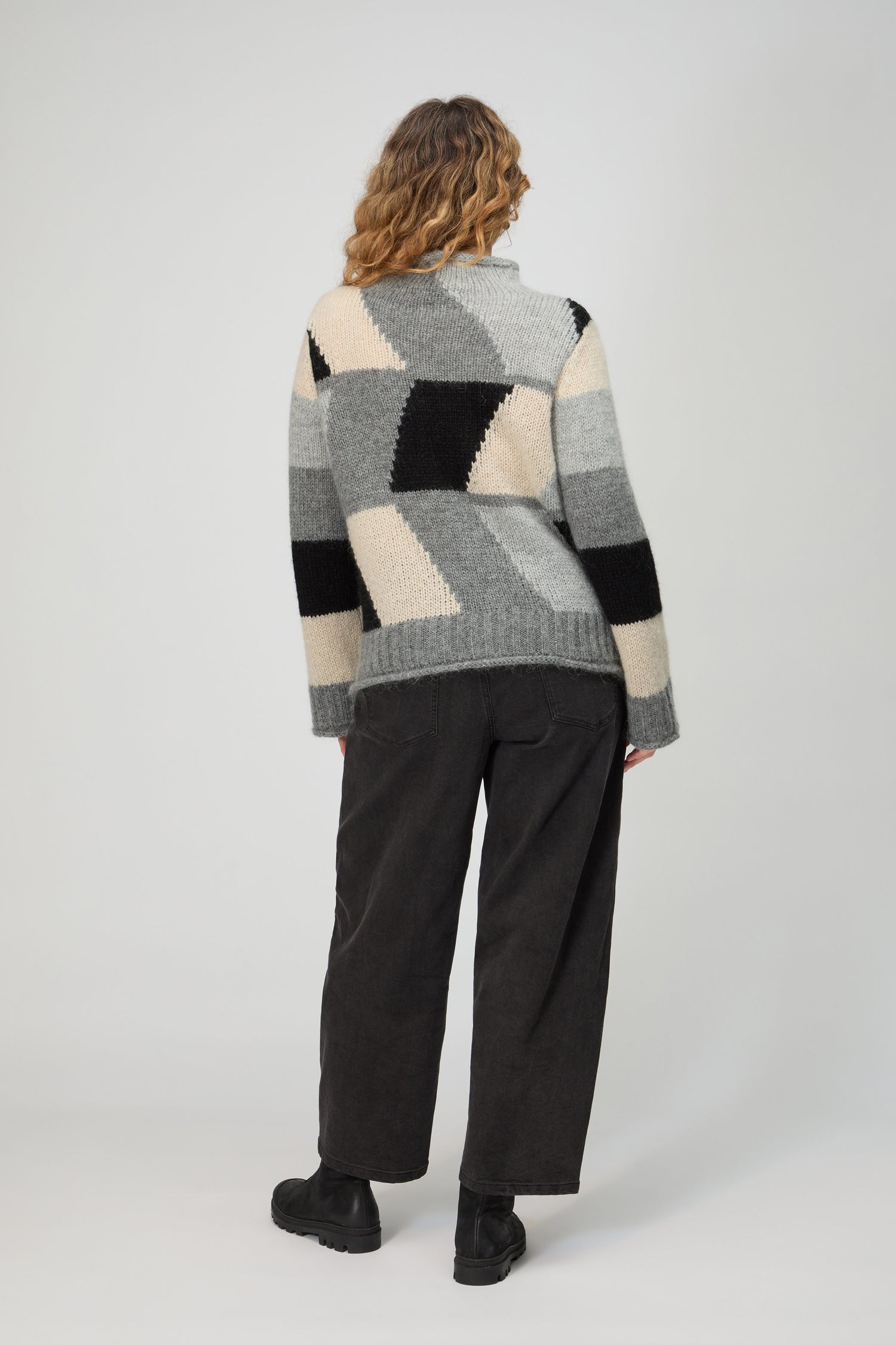 A person stands facing away in the Grey Colour Block Knit—shades of grey, black, and cream—paired with dark pants and brown shoes against a plain background, channeling effortless Scandinavian minimalism.