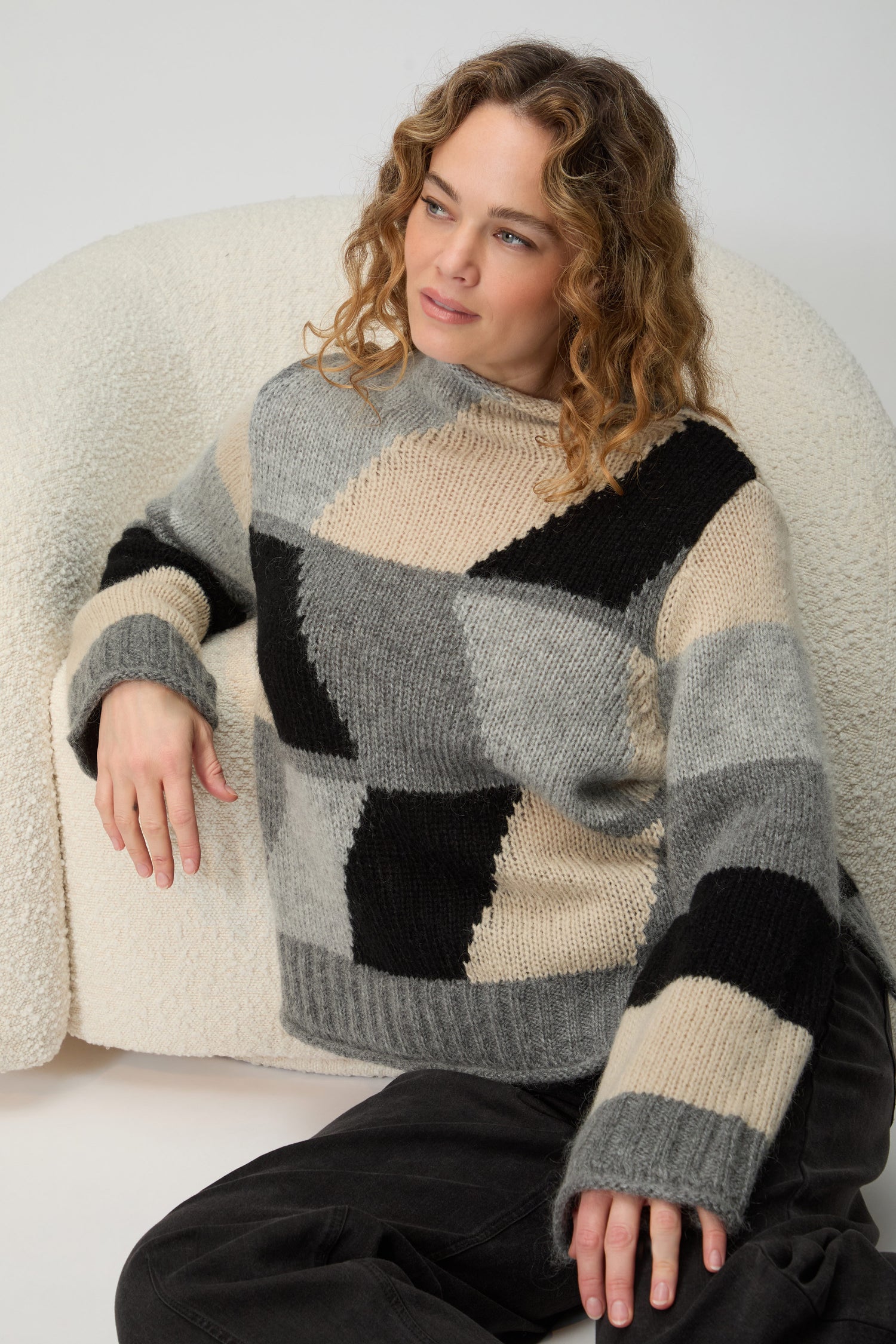 A woman with long brown hair wears the Grey Colour Block Knit in gray, black, and beige with dark pants, standing against a plain light background for a Scandinavian minimalist look.