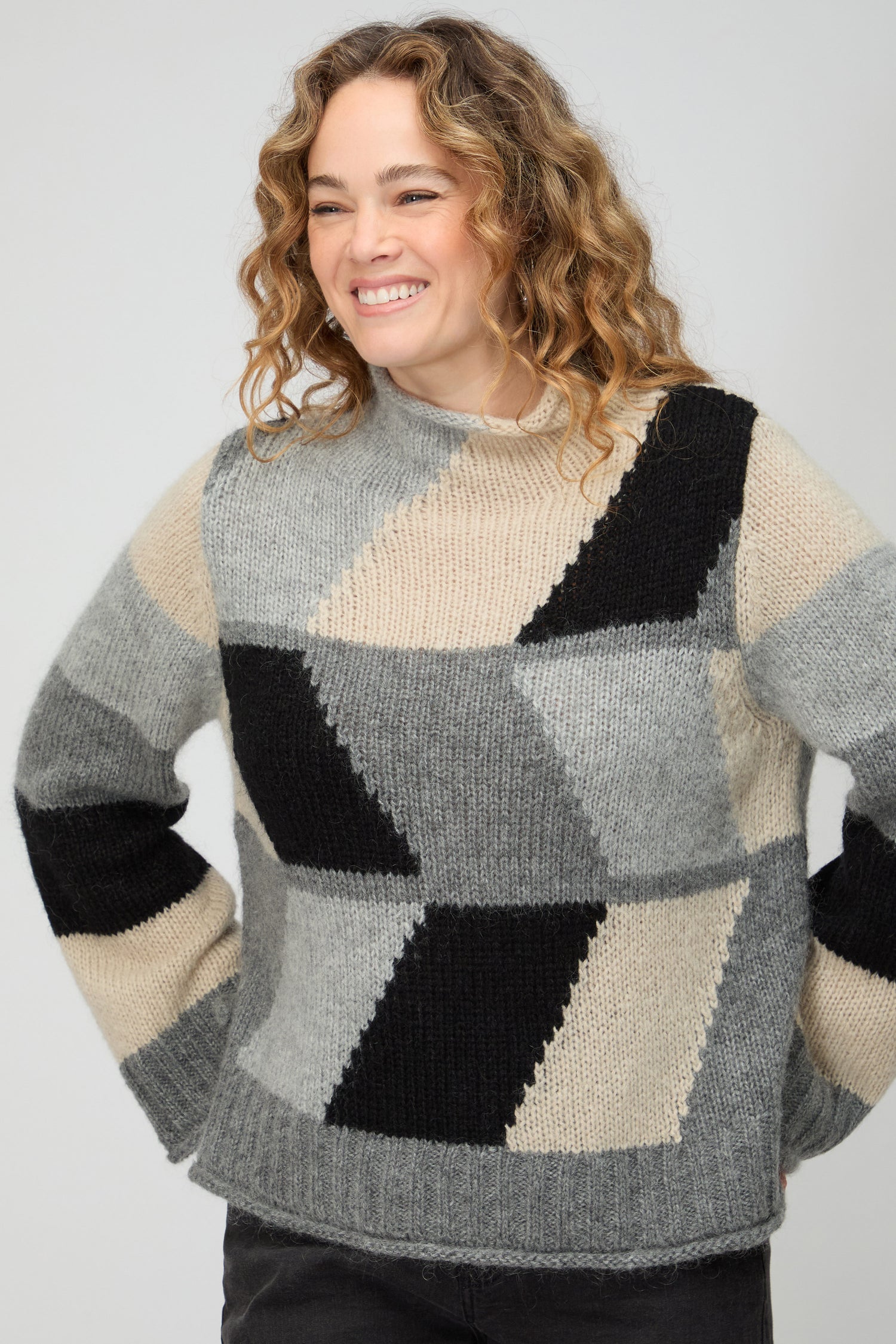 A woman stands against a plain background wearing the Grey Colour Block Knit, loose dark pants, hoop earrings, and beige shoes—an effortless nod to Scandinavian minimalism.