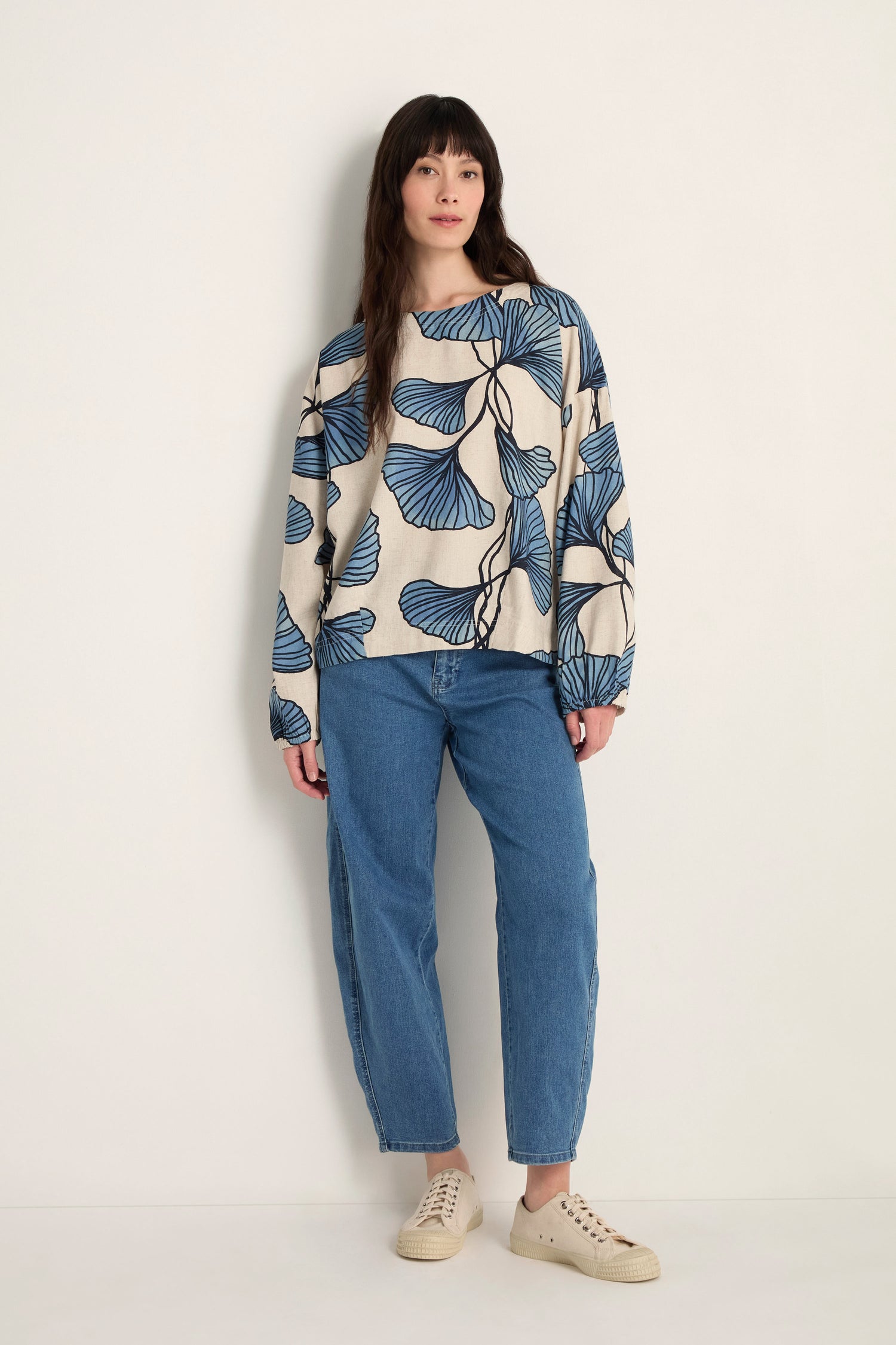 A person stands against a plain wall wearing a Ginkgo Viscose Linen Boxy Top with a blue leaf pattern, blue jeans, and beige sneakers.