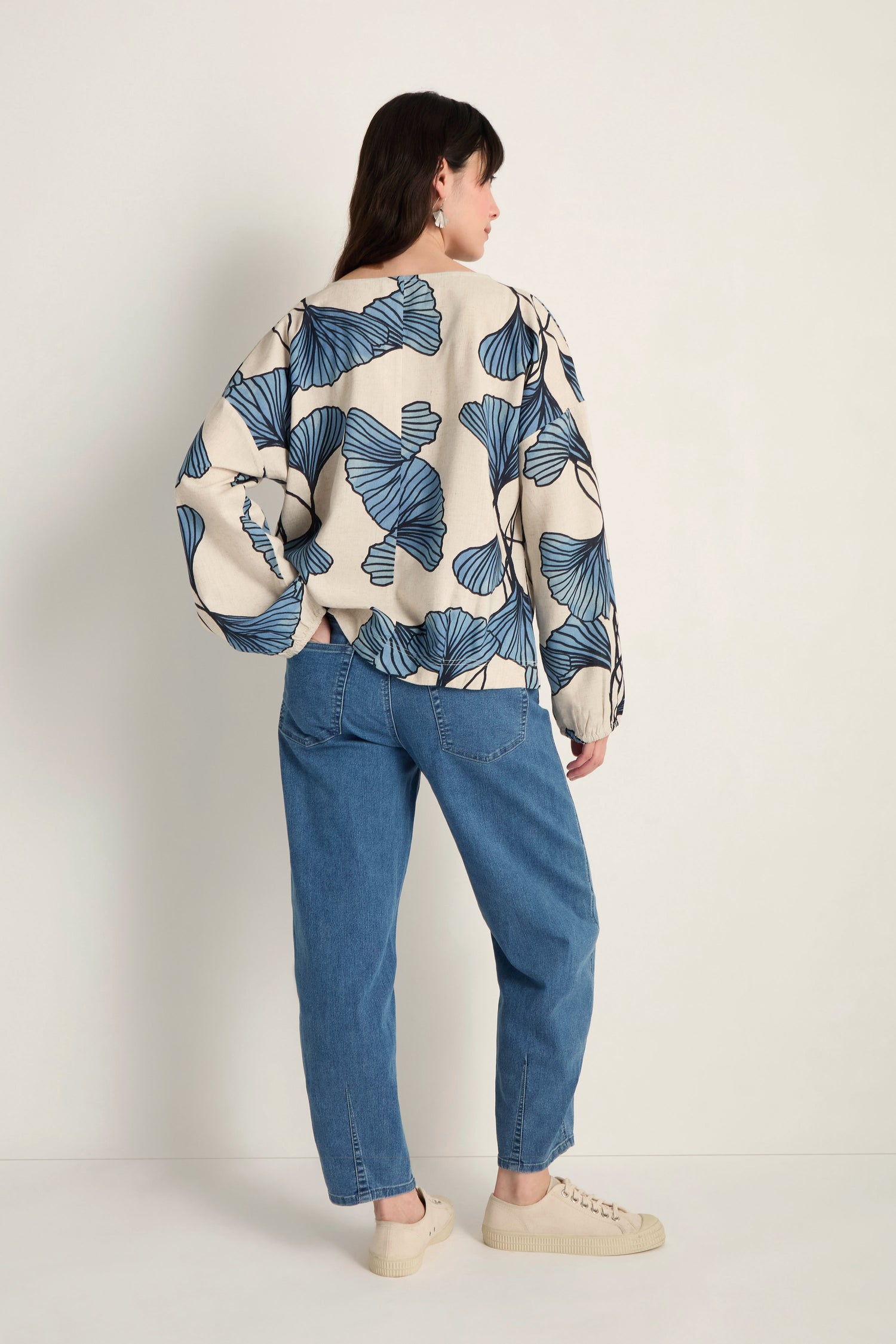 A person stands facing away, wearing a Ginkgo Viscose Linen Boxy Top with a blue leaf pattern, blue jeans, and beige shoes against a plain background.