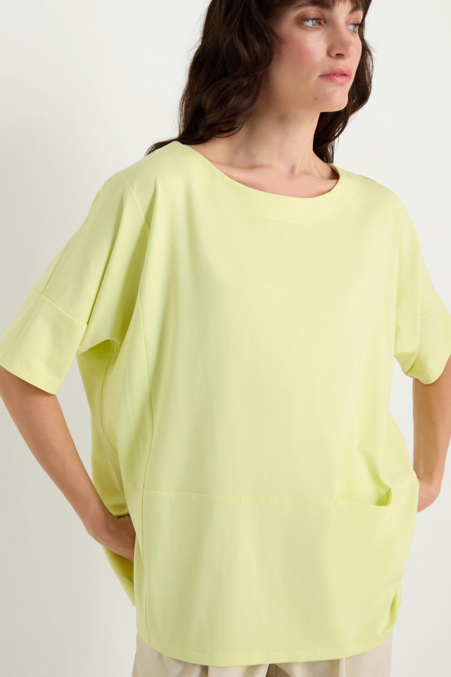 A woman wears the Jersey Pocket Top, a relaxed fit light yellow viscose jersey with wide sleeves and a front pocket, paired with beige pants, standing against a plain light background.