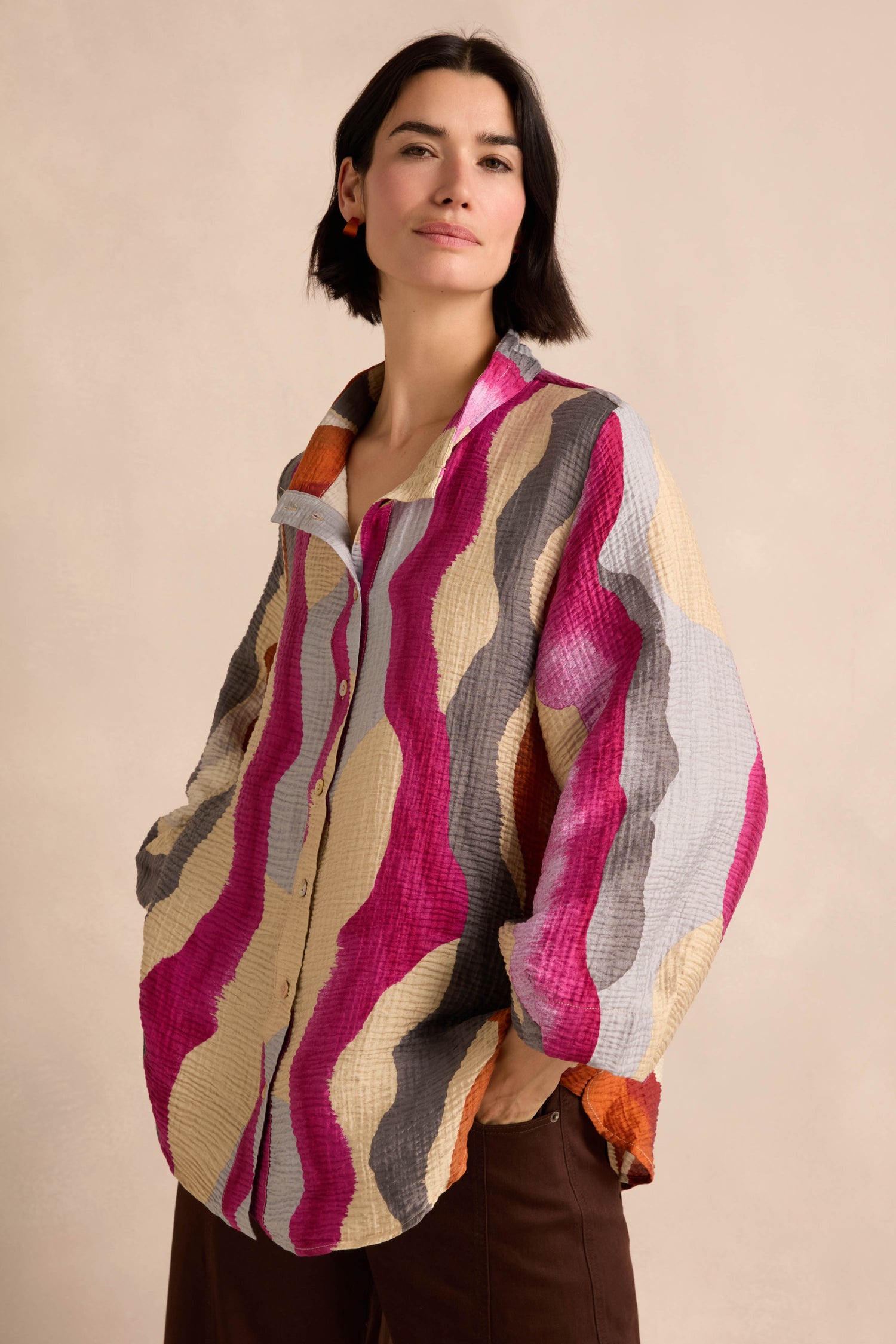 A person wearing the Wavy Painted Lines Shirt—a loose, button-up crinkled viscose shirt featuring wavy pink, beige, gray, and orange stripes—stands with hands in pockets against a neutral background.