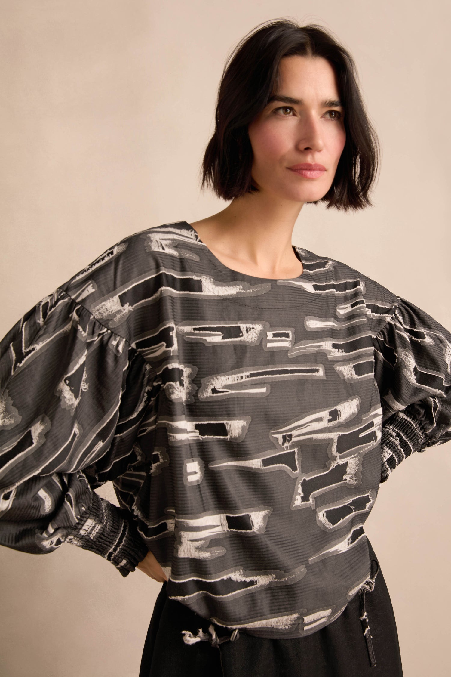 A woman with short dark hair models the Abstract Embroidery Top, a loose, long-sleeve black and gray patterned blouse featuring abstract embroidered details, paired with black pants. She stands with her hands on her hips against a neutral background.