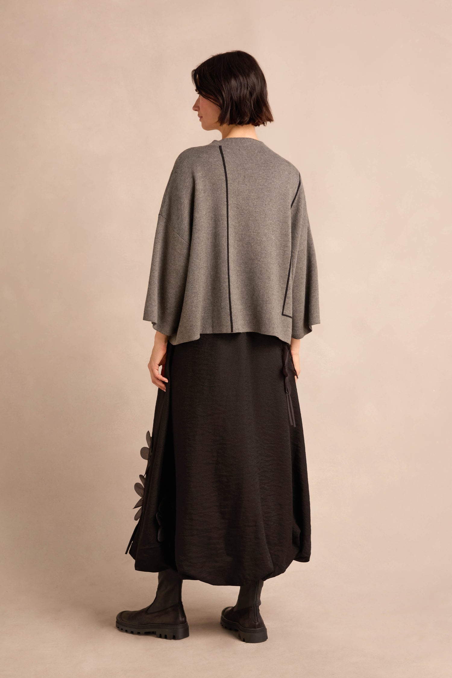 A person stands with their back to the camera, wearing the Piping Detail Knit—a relaxed fit gray top—paired with a long black skirt featuring textured details and black boots, set against a neutral background.