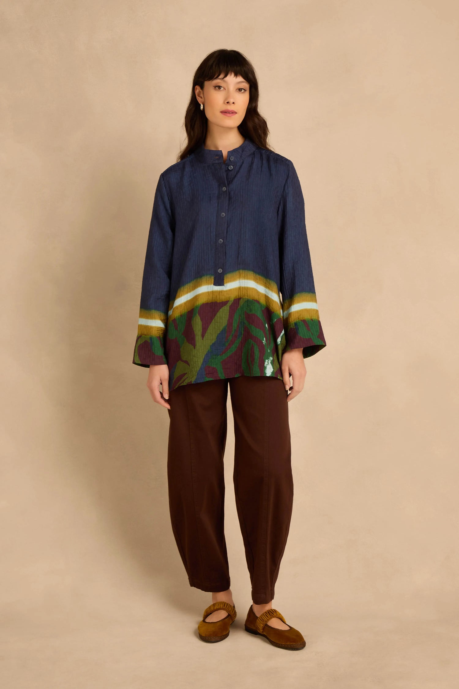 A woman wears a relaxed A-line Print Hem Pocket Shirt in dark blue with a multicolored abstract print hem, paired with brown trousers and olive flats.