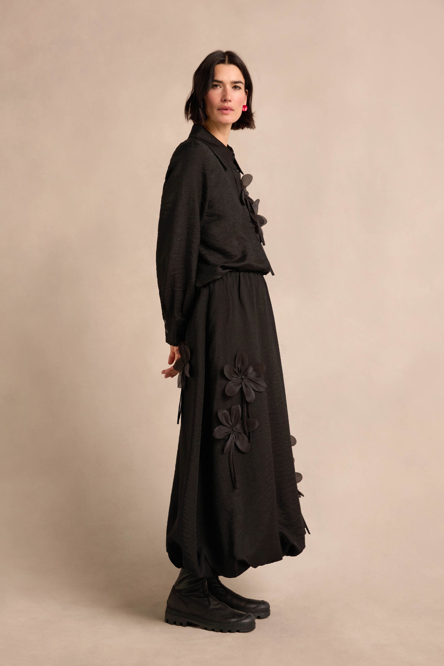 A person standing against a neutral background, wearing the Flower Detail Bubble Skirt and a matching black top with delicate floral appliqué details, paired with black boots.