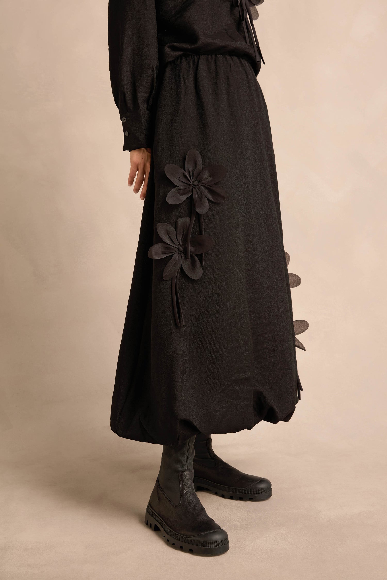 A person wearing the Flower Detail Bubble Skirt—a long black midi skirt featuring large floral appliqués—paired with black boots, standing against a neutral background.