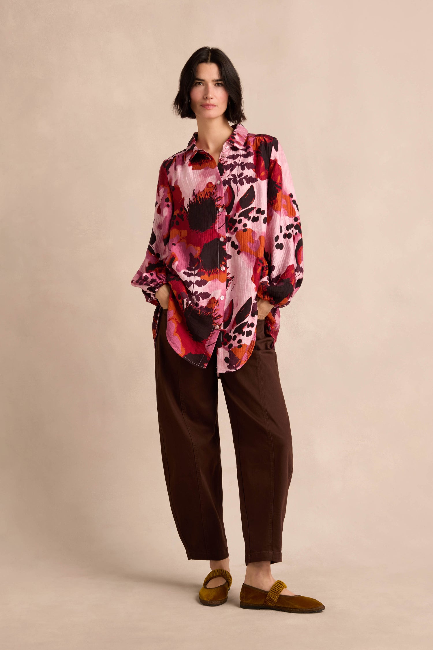 A person stands against a plain background wearing the Crinkle Viscose Shirt in a loose pink botanical print, paired with brown pants and brown slip-on shoes, with hands in pockets.