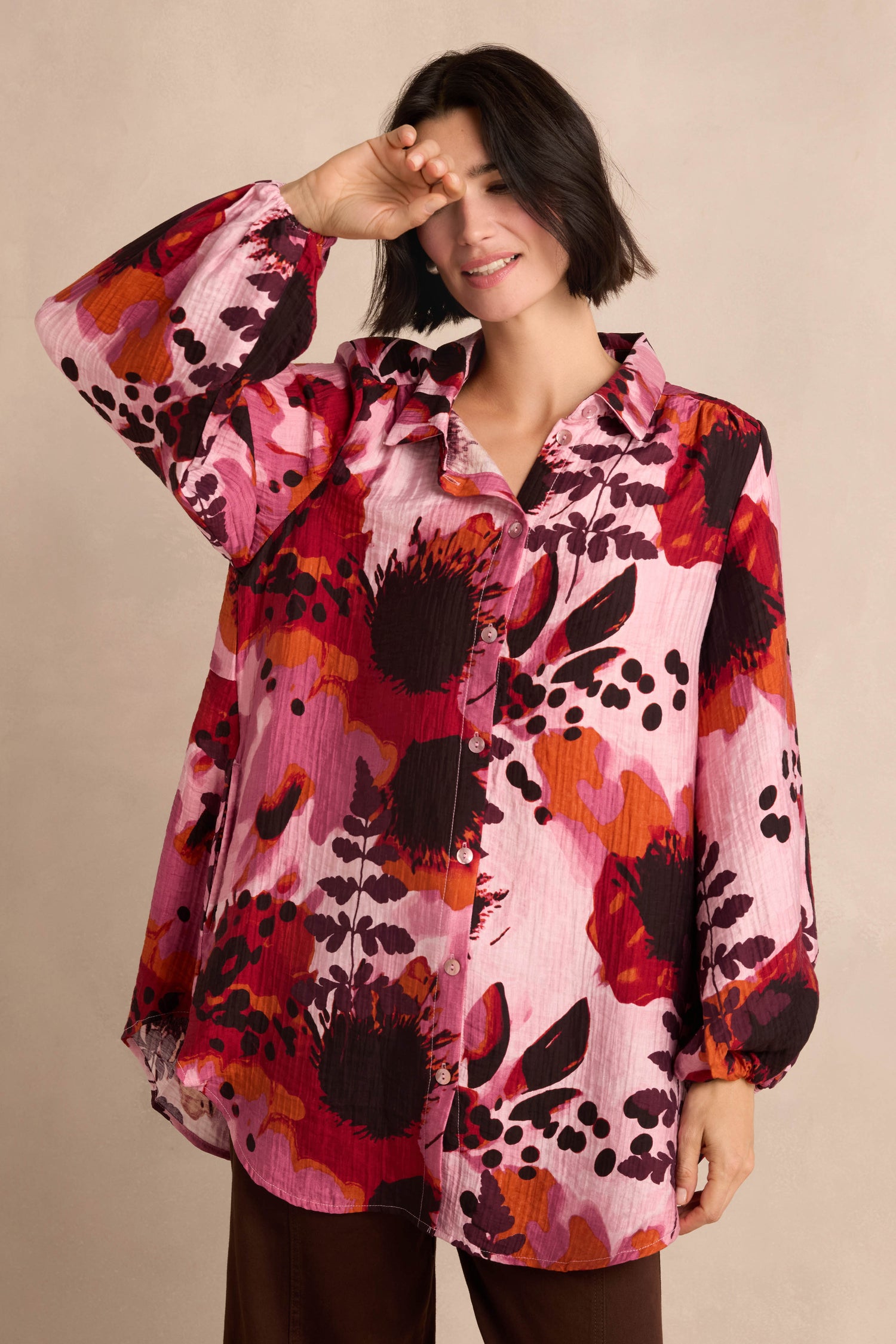 A woman stands against a beige background, smiling and shielding her eyes with one hand, wearing the Crinkle Viscose Shirt—a loose, long-sleeve blouse in pink and red botanical print—paired with brown pants.
