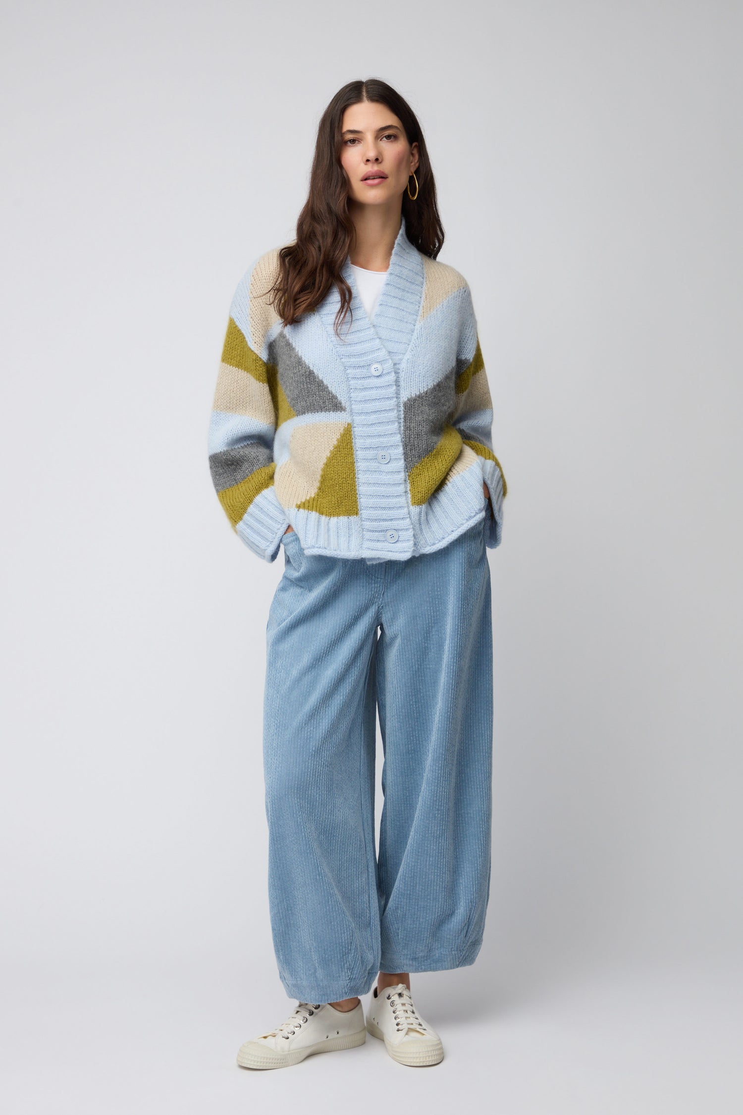 A woman stands against a plain background wearing the Colour Block Cardi, wide-leg blue pants, white shoes, and hoop earrings.