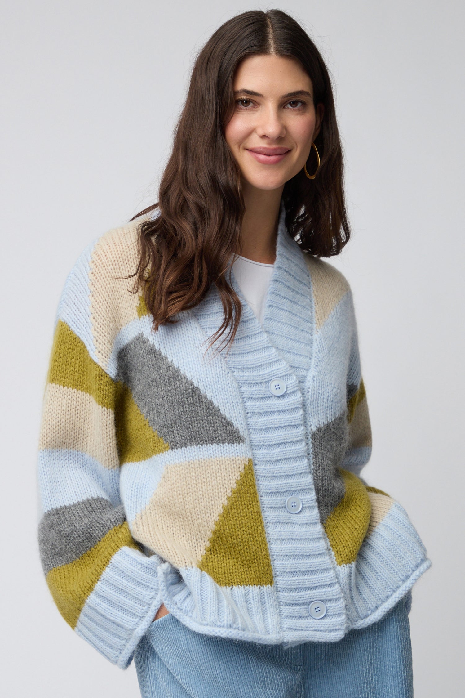 A woman with long brown hair wears the Colour Block Cardi in pastel blue, olive, cream, and gray with light blue pants, standing against a plain background.