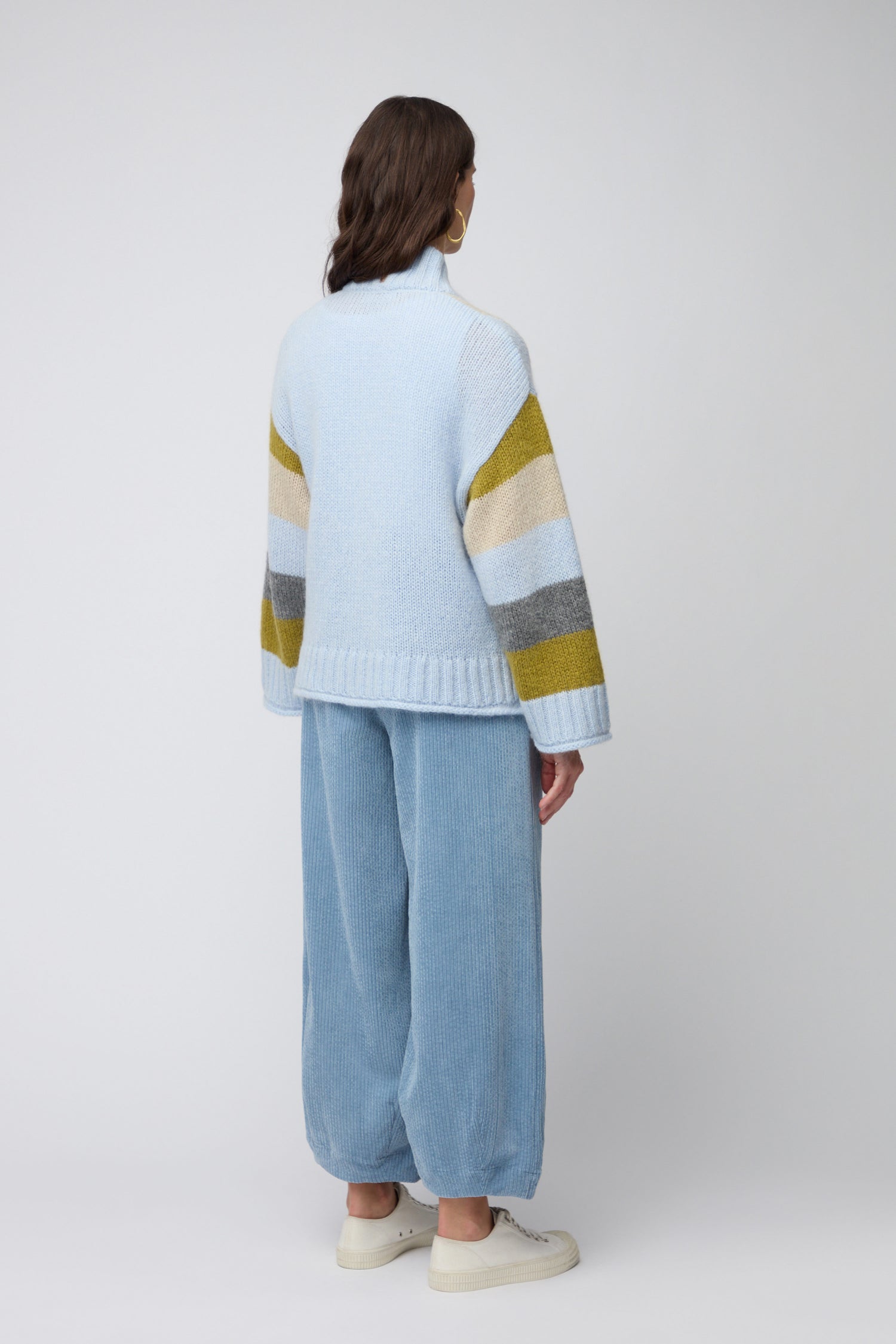 A person faces away, wearing the Colour Block Cardi—a light blue contemporary knitwear sweater with striped sleeves—paired with loose blue corduroy pants and white sneakers, set against a neutral background.