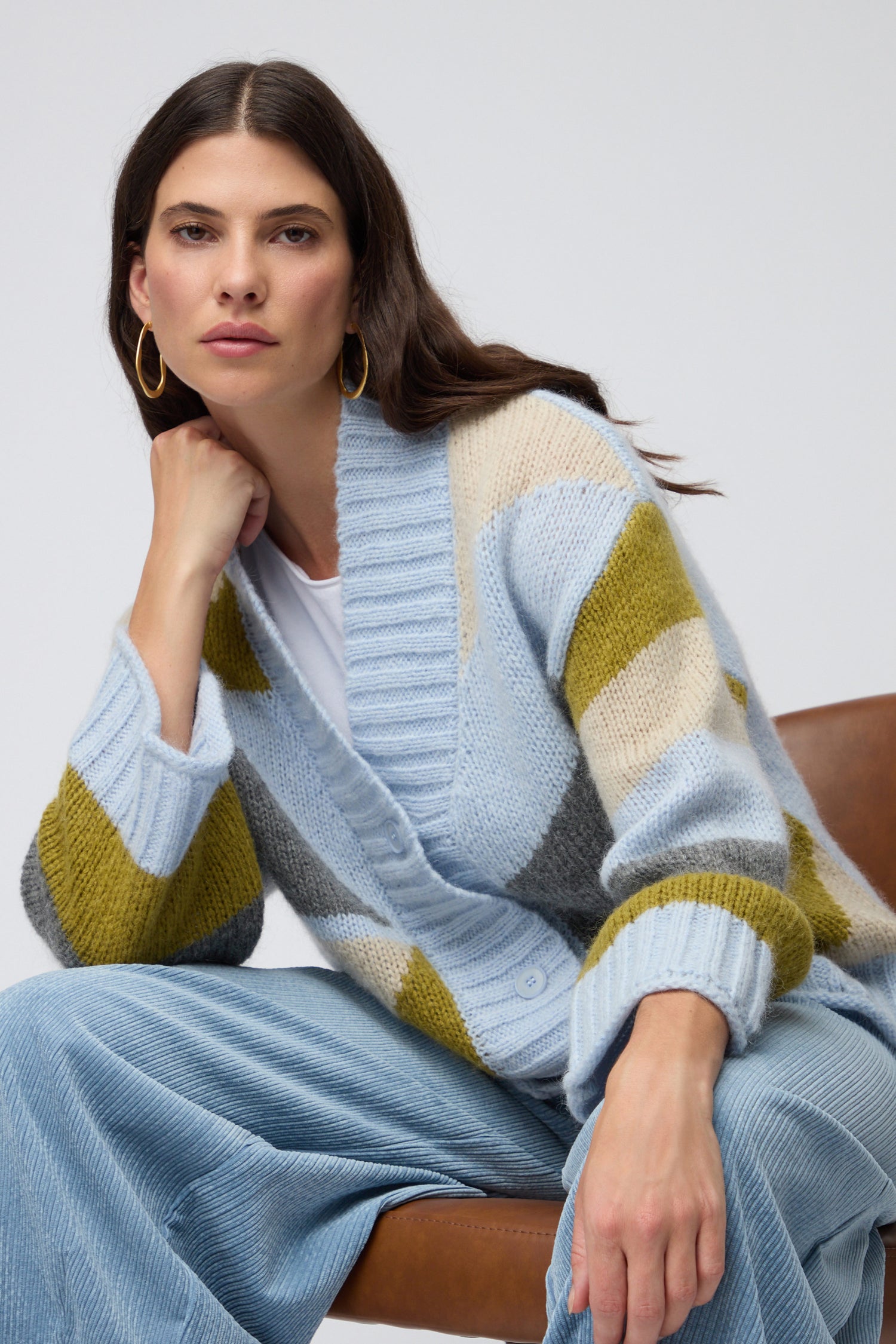 A woman with long brown hair sits on a chair wearing the Colour Block Cardi—blue, gray, and yellow—from a Scandinavian brand, paired with a white shirt, blue pants, and gold hoop earrings, looking at the camera.