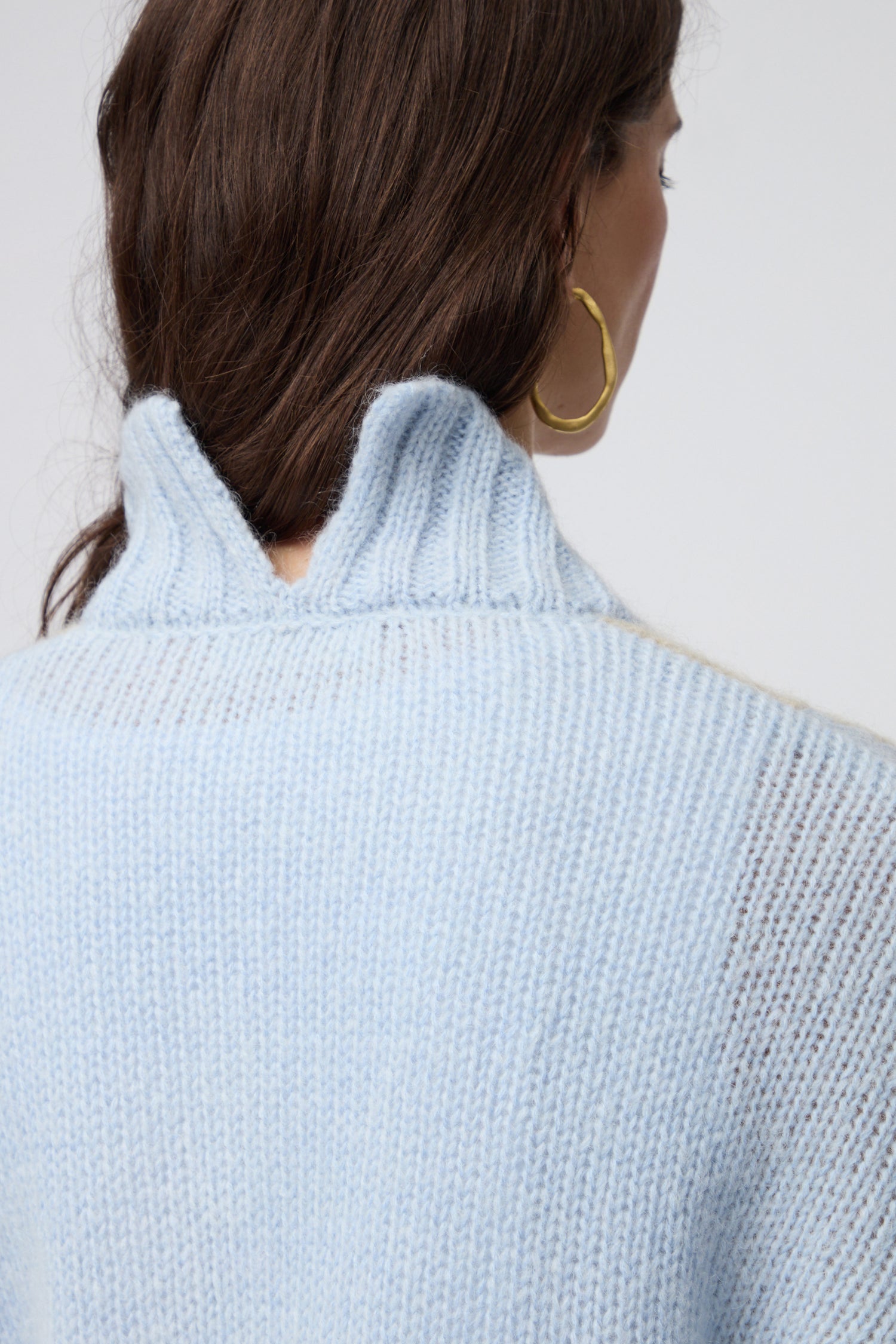 A person with long brown hair, seen from behind, wears the Colour Block Cardi—a light blue ribbed knit sweater with a wavy collar—styled with large gold hoop earrings for a sleek Scandinavian-inspired look.