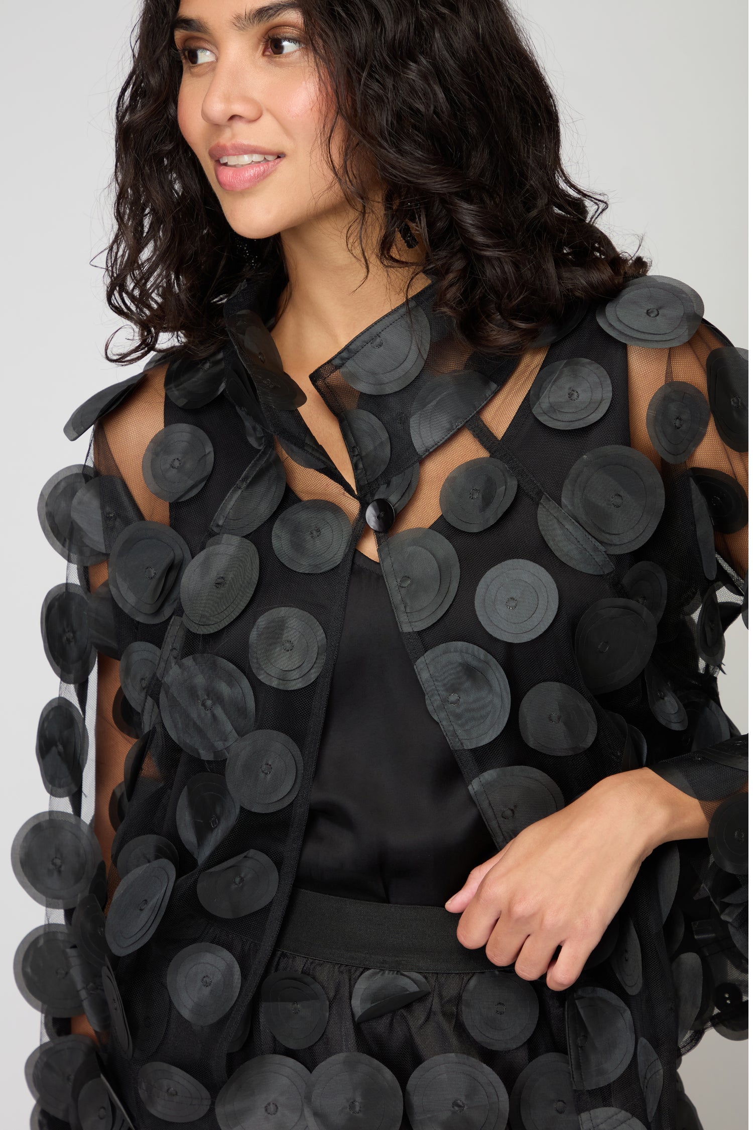 A woman models the Cut Out Circles Jacket, a sheer black piece inspired by Scandinavian fashion, standing against a plain light background.