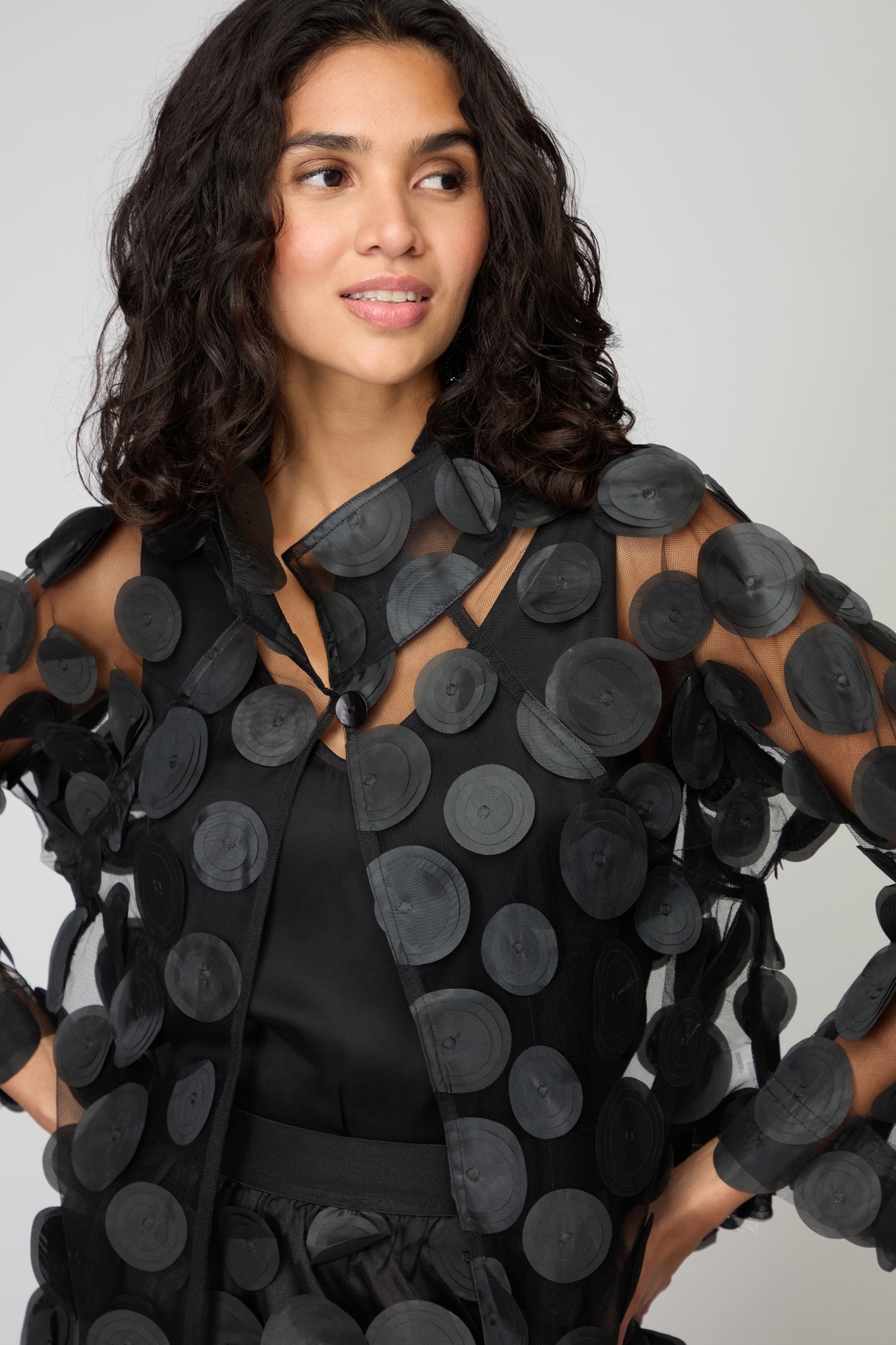 A woman with wavy dark hair models the Cut Out Circles Jacket—black mesh with bold circular appliqués—showcasing sleek Scandinavian style against a simple light background.