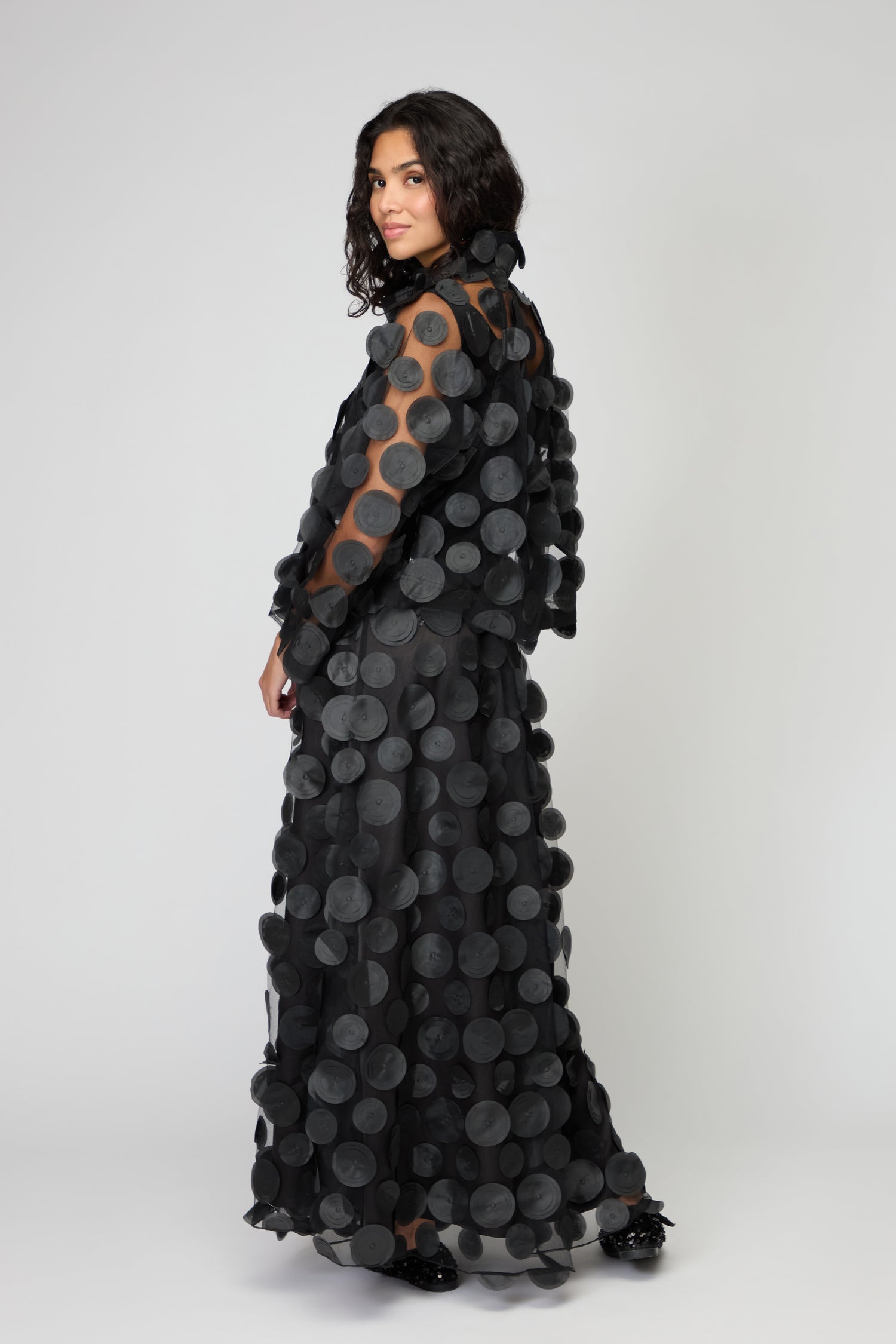 A woman in the Cut Out Circles Jacket, featuring bold circular appliqués, stands against a plain light background, glancing over her shoulder.